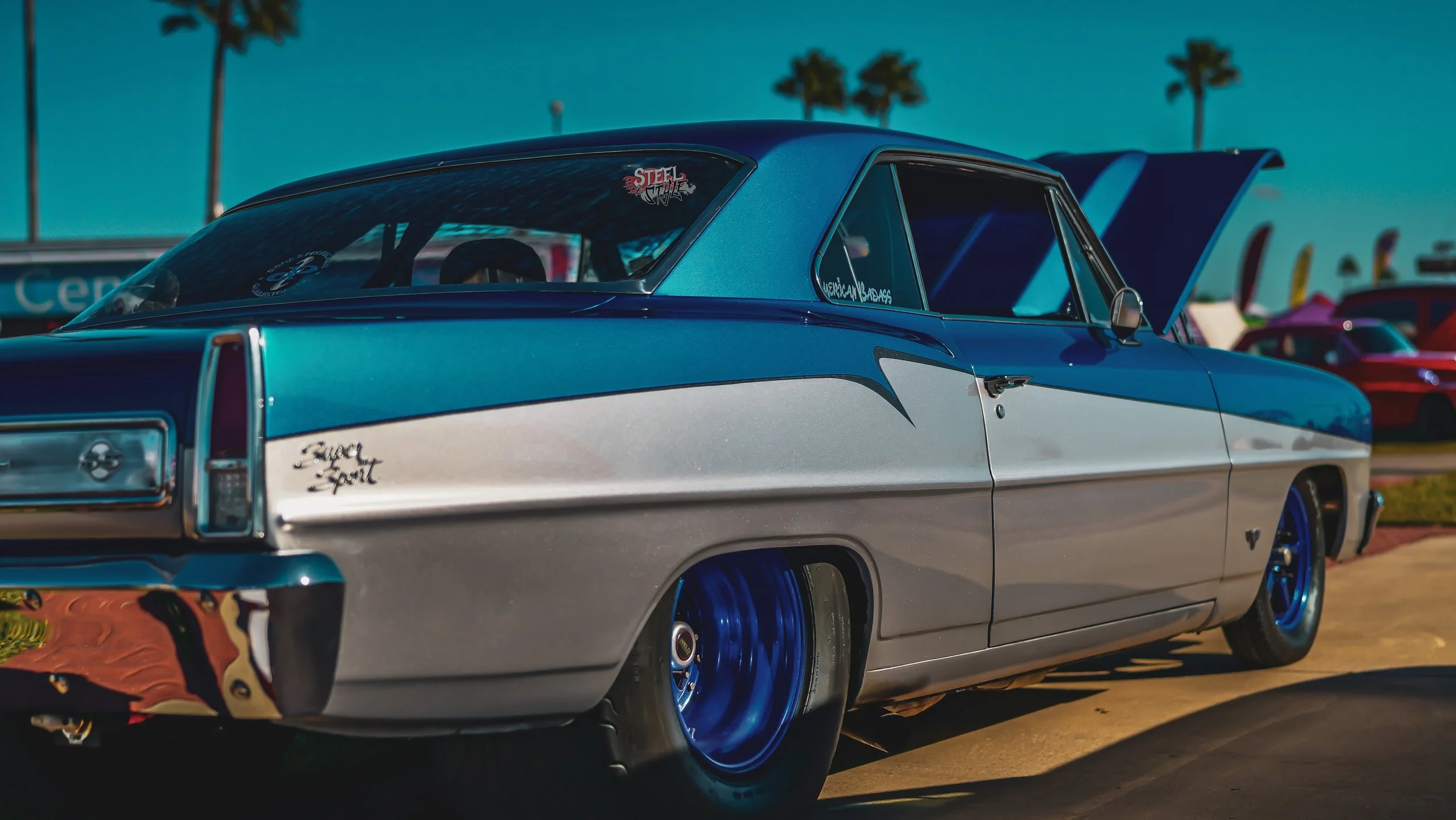 A vintage drag racing car with a two-tone blue and silver paint job, lowered stance, and blue wheels, parked at a racing event with other cars and palm trees in the background.