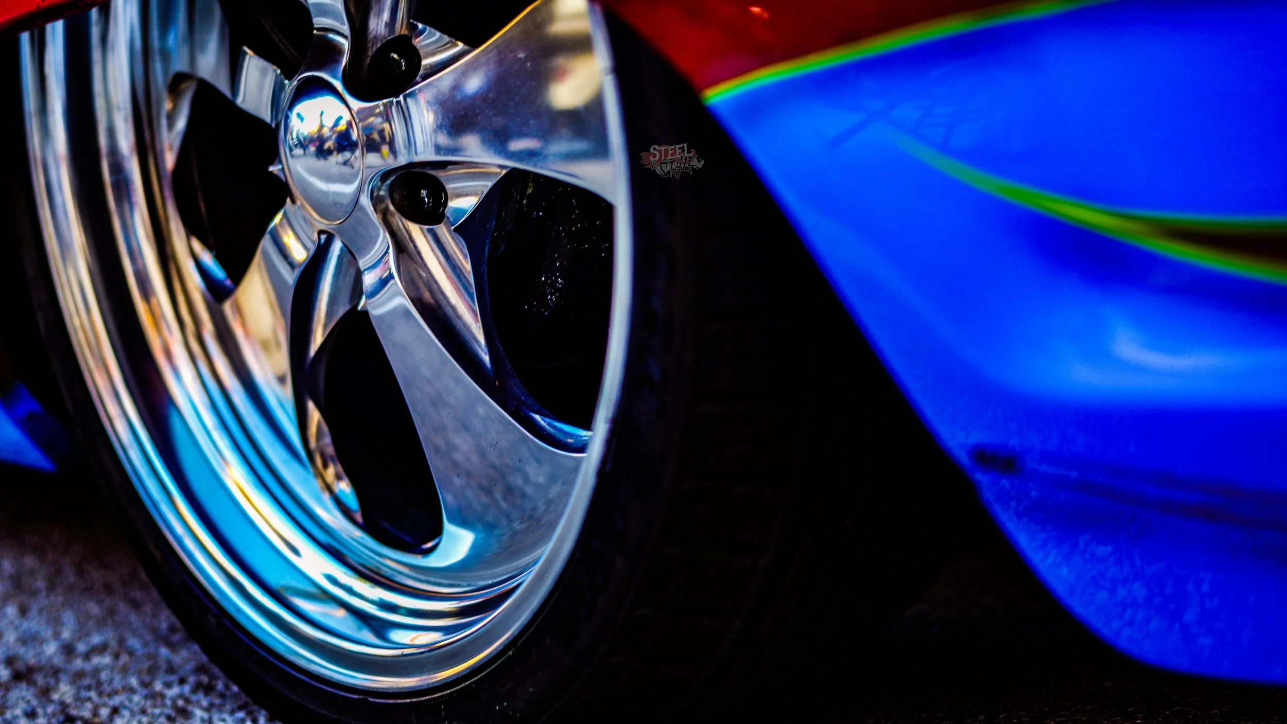 Close-up of a polished chrome car wheel and part of a blue car body in an outdoor setting.