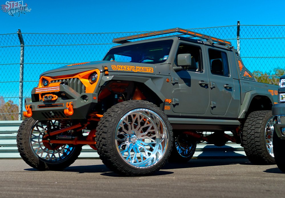 A modified off-road pickup truck with elevated suspension, large chrome wheels, gray body with orange accents, custom bumper, and roof rack, parked at a racetrack.