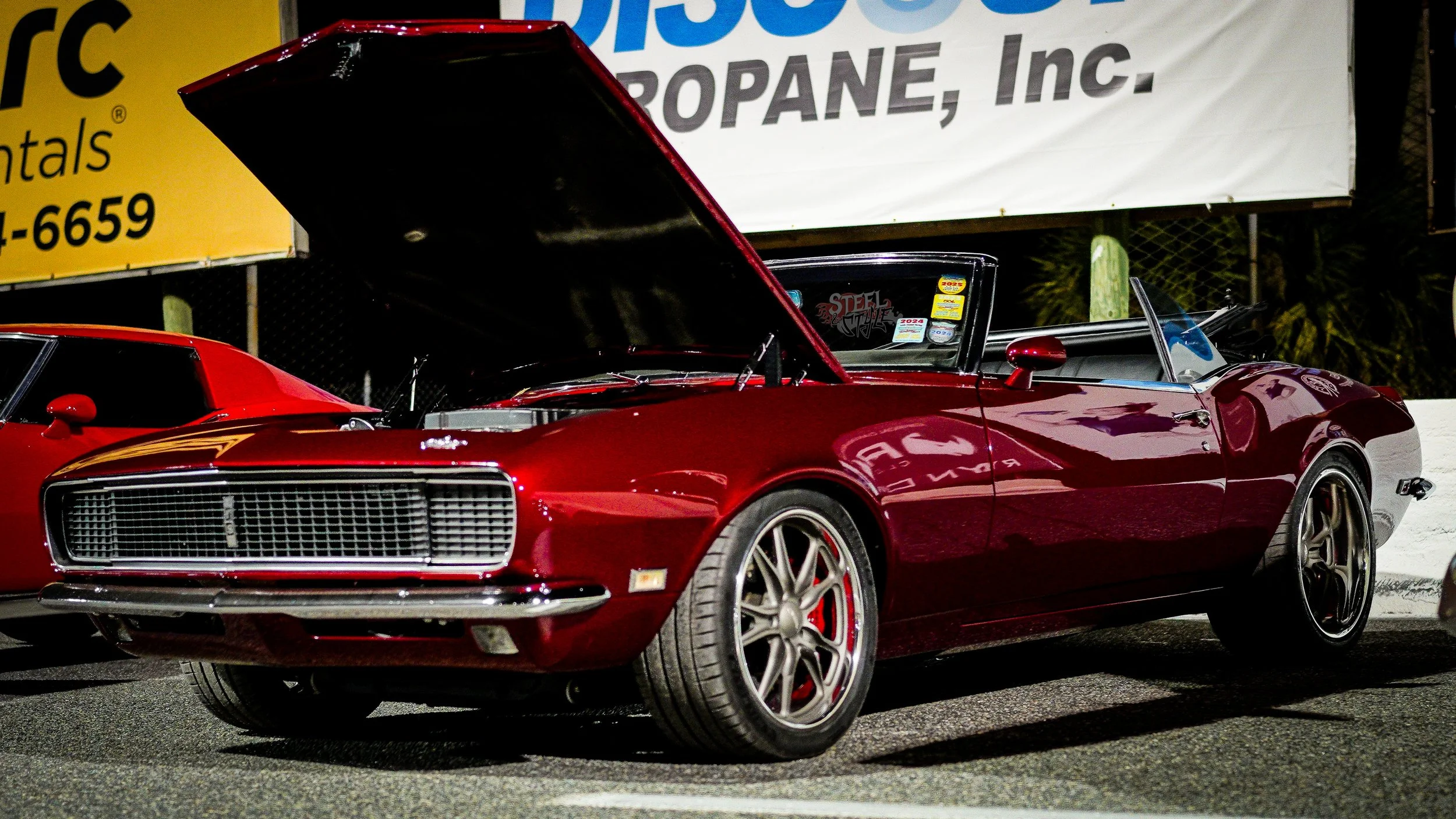 A shiny red vintage sports car with its hood open, parked next to another red car at a nighttime event.