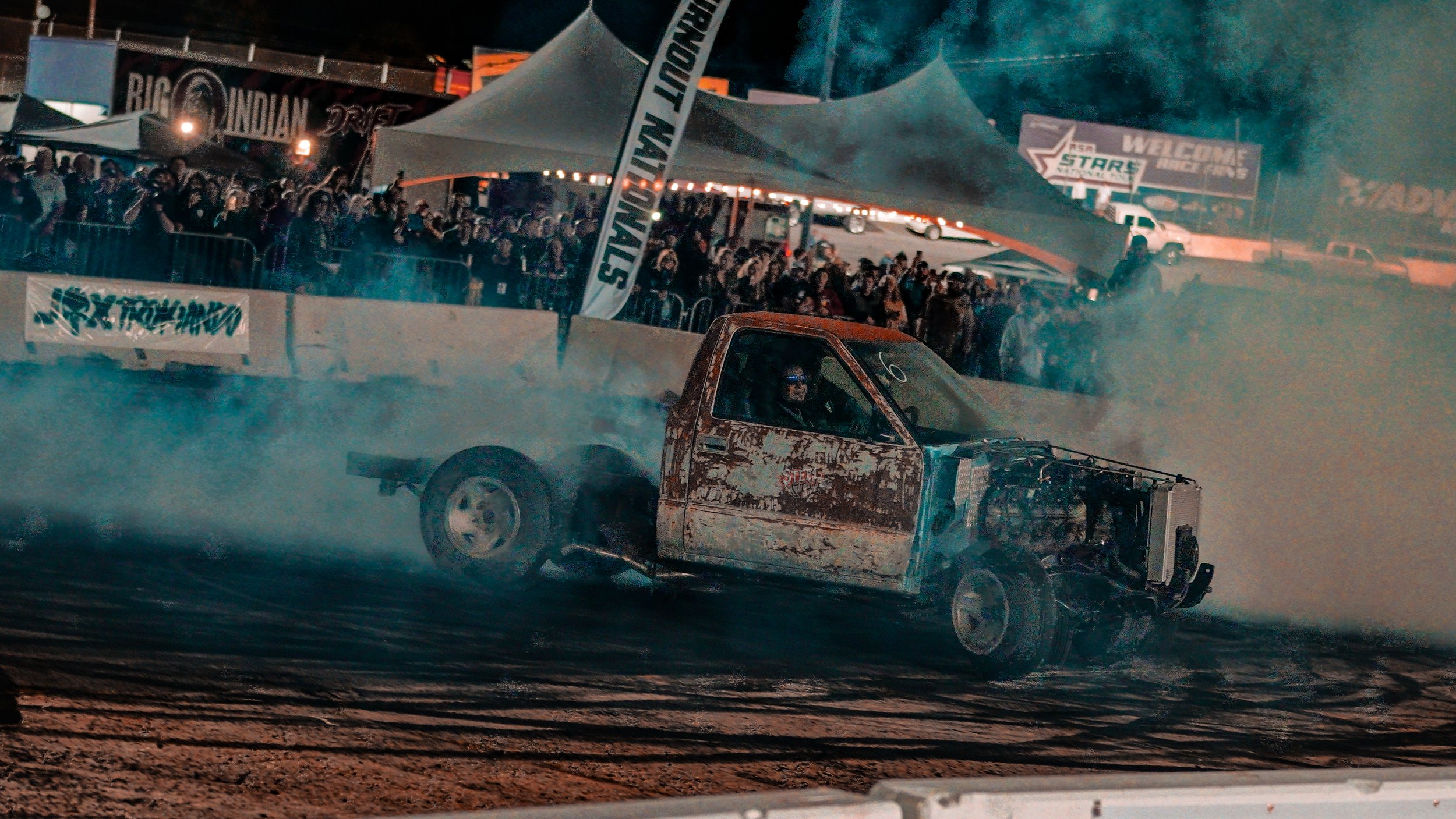 A rusty, old pickup truck drifting on a dirt track during a car racing event at night with spectators watching behind barriers and tents.