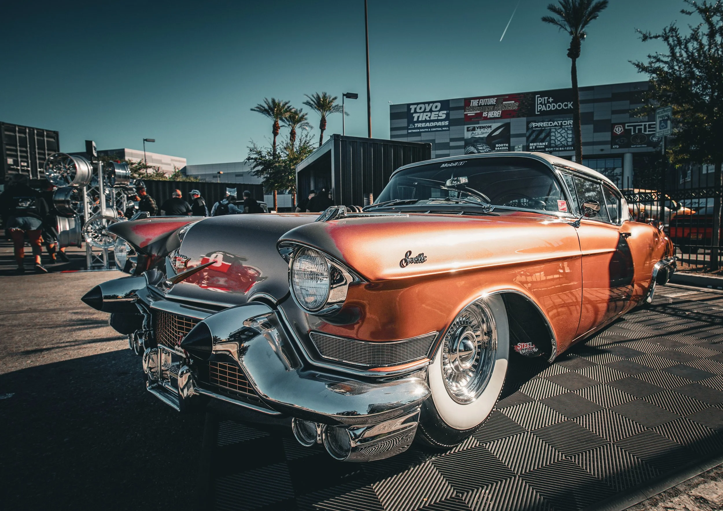 A vintage gold and silver Cadillac car on display at a car show, with palm trees and commercial buildings in the background.