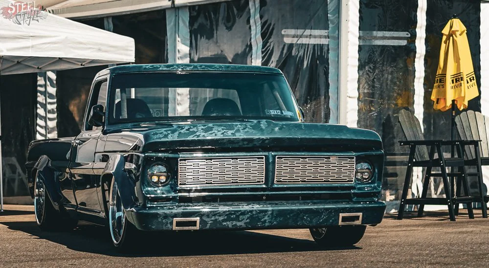 A black vintage pickup truck with bold custom grille and lowered stance parked outdoors near a white canopy and yellow umbrella.