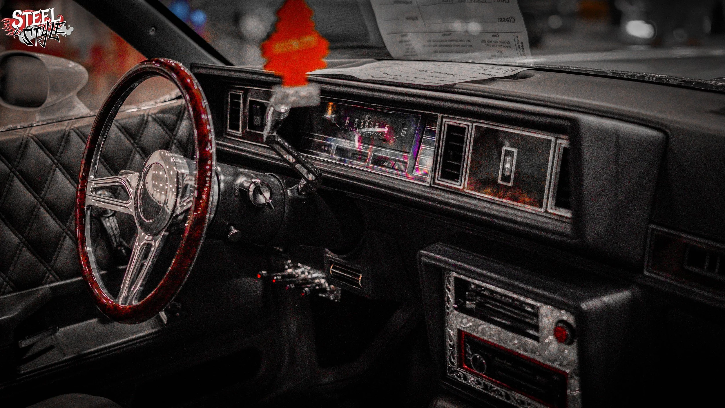 The interior of a vintage car with a custom dashboard, diamond-stitched black leather seats, a shiny red and chrome steering wheel, and an air freshener hanging from the rearview mirror.
