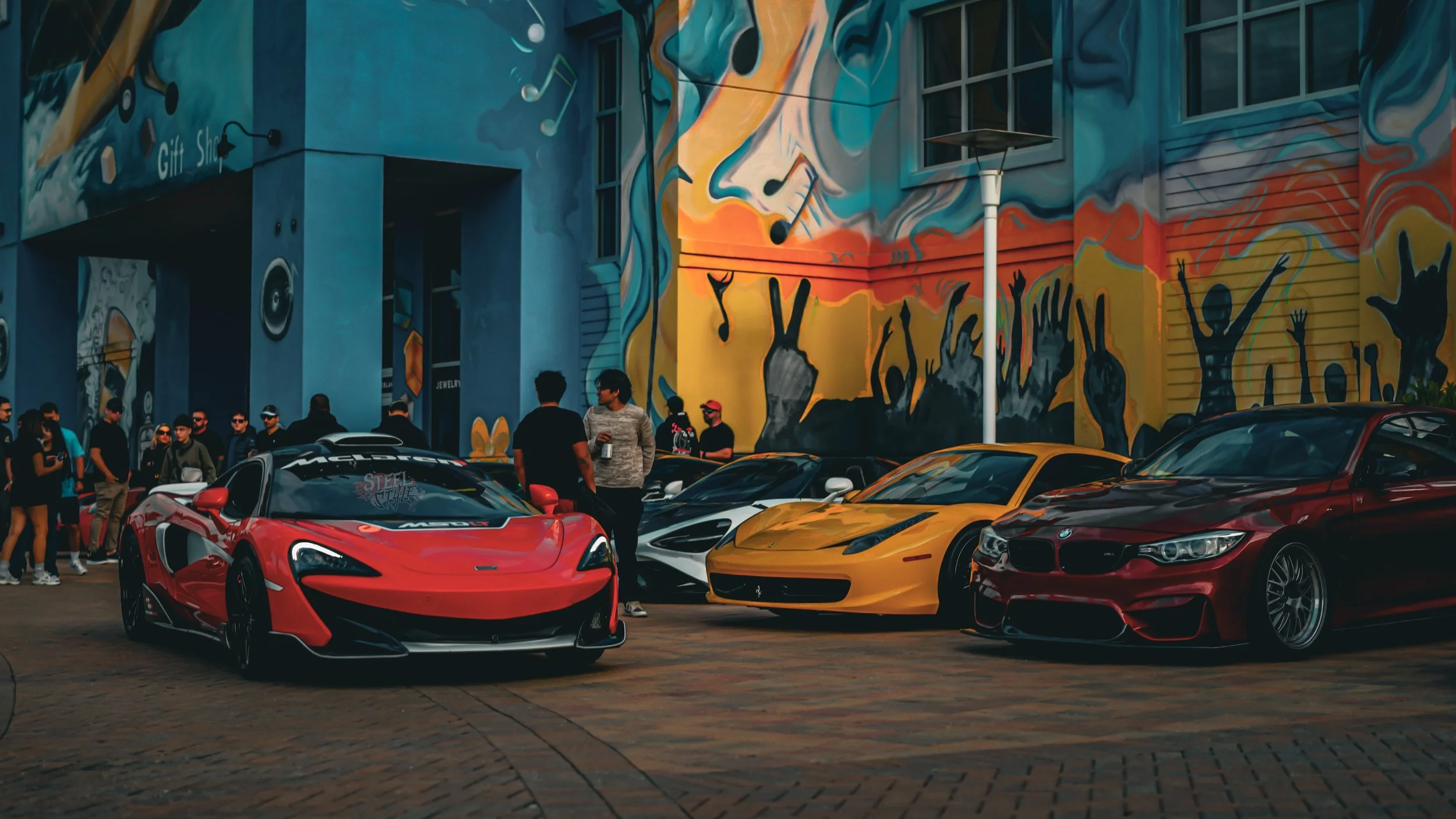A lineup of luxury sports cars parked in front of a colorful mural with silhouettes of people and musical notes, with a group of people standing nearby.