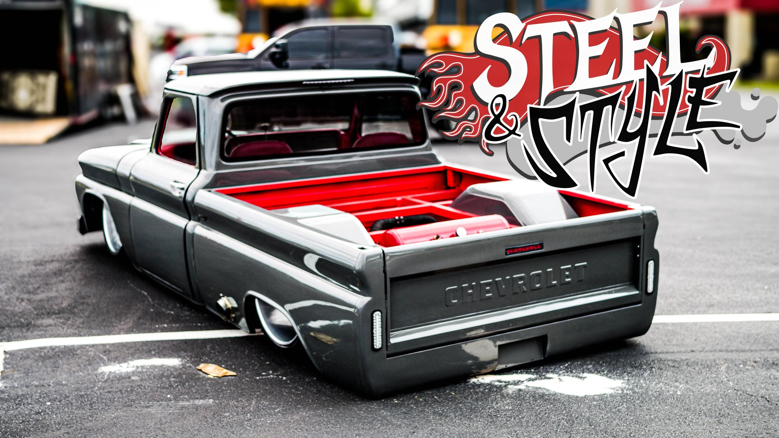 A custom low-rider pickup truck with a black and gray paint job and red interior, parked in a lot. graphics with the words 'Steam & Style' are overlaid on the upper right side.