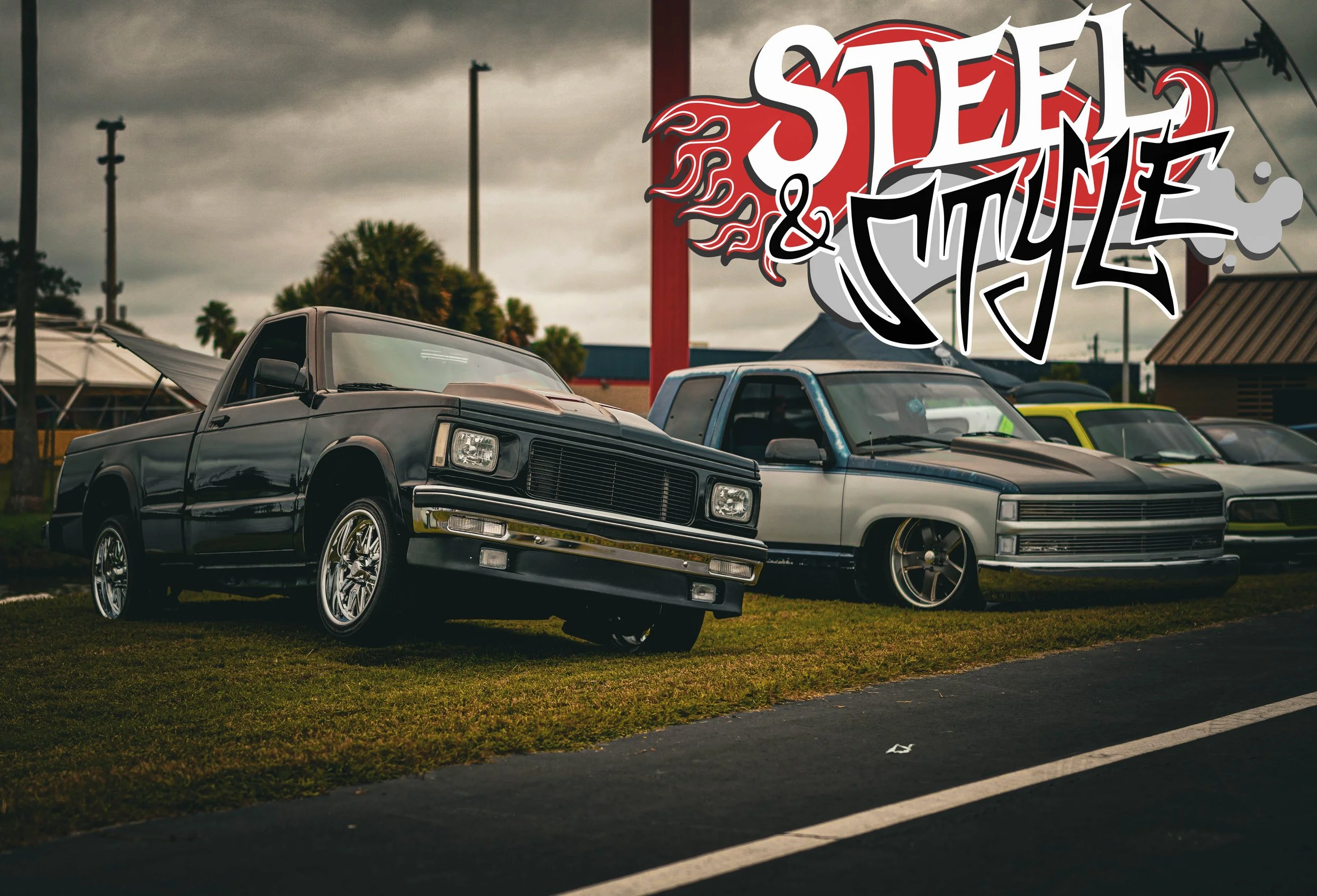 A lineup of customized classic cars on a grassy area with a sign that reads 'Steel & Style' in graffiti font, under an overcast sky.
