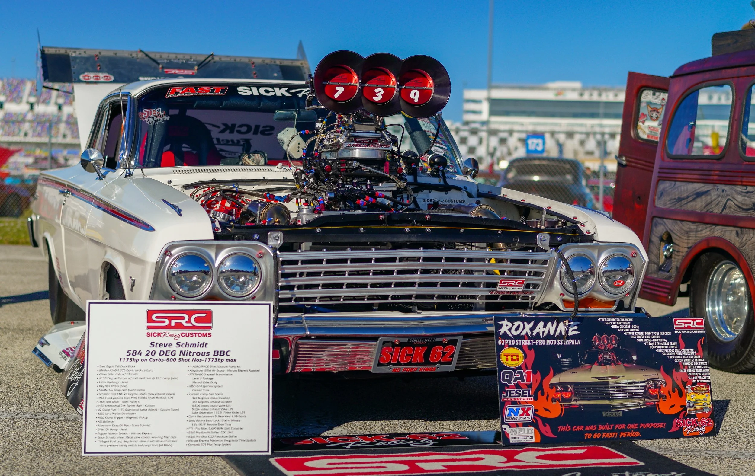 Classic race car with exposed engine on display at a racing event, with informational signs and other vintage vehicles in the background.