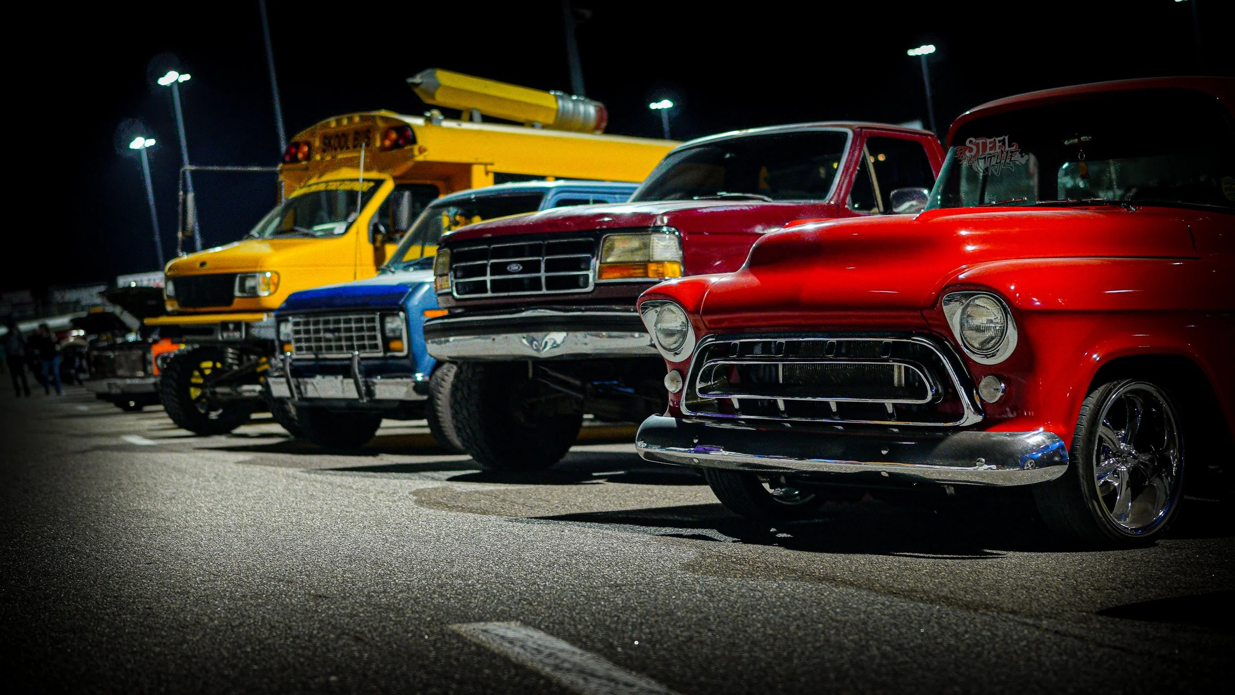A lineup of vintage cars parked in a lot at night, including a red classic automobile in the foreground, a blue truck, and a yellow utility vehicle with a rocket in the background, illuminated by overhead parking lot lights.