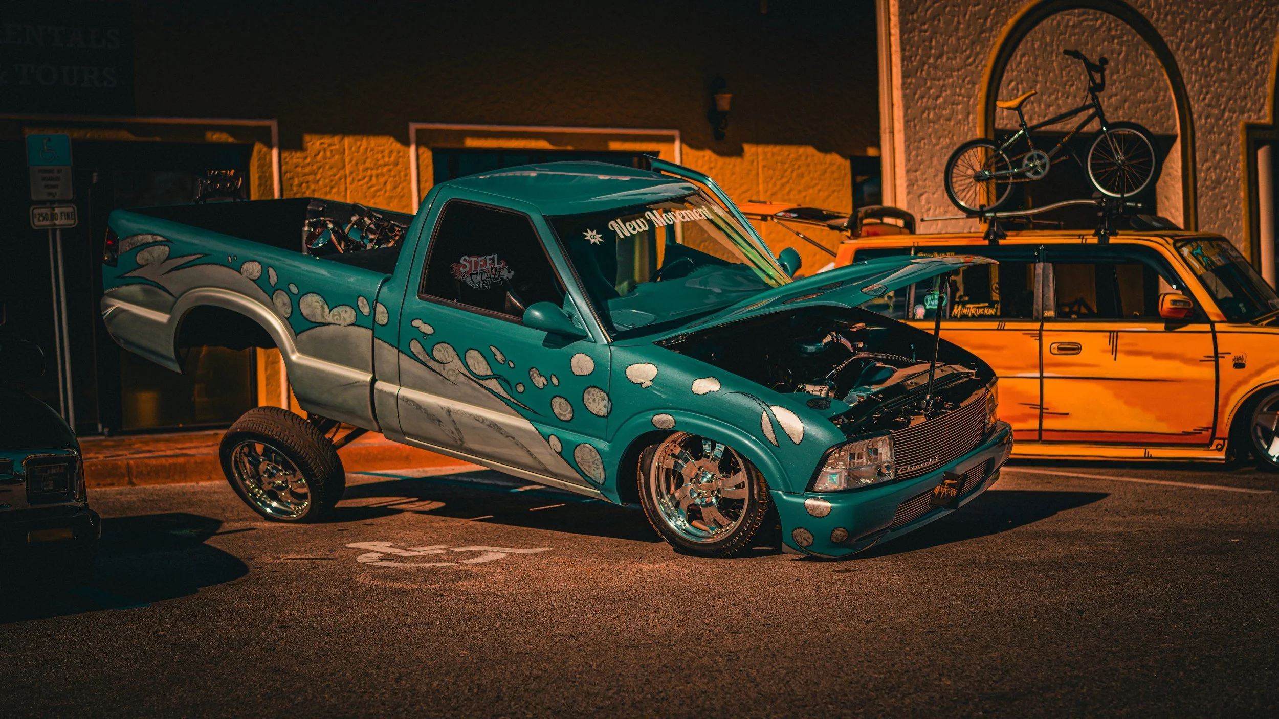 A customized teal pickup truck with an open hood displaying an engine, parked in a lot at night. The truck has silver flames and bubbles painted on its side, large shiny chrome wheels, and a sticker on the windshield that reads "New Movement." Behind