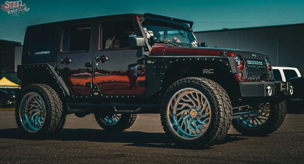 A custom red and black off-road vehicle with large tires, alloy wheels, and aftermarket modifications, parked outdoors during the daytime.