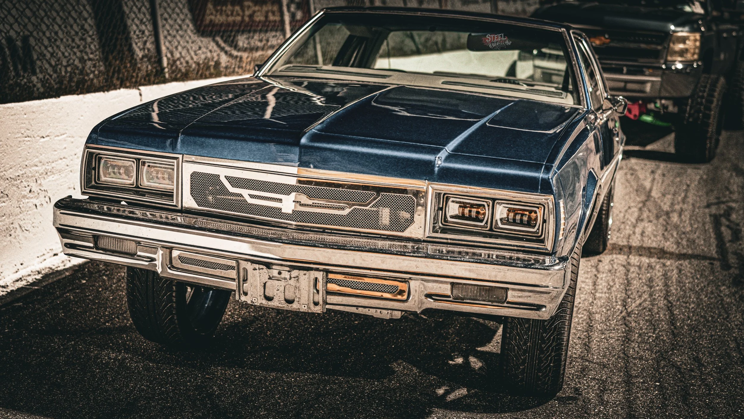 A vintage blue Chevrolet Malibu car parked on the street at night, with another large vehicle visible in the background.