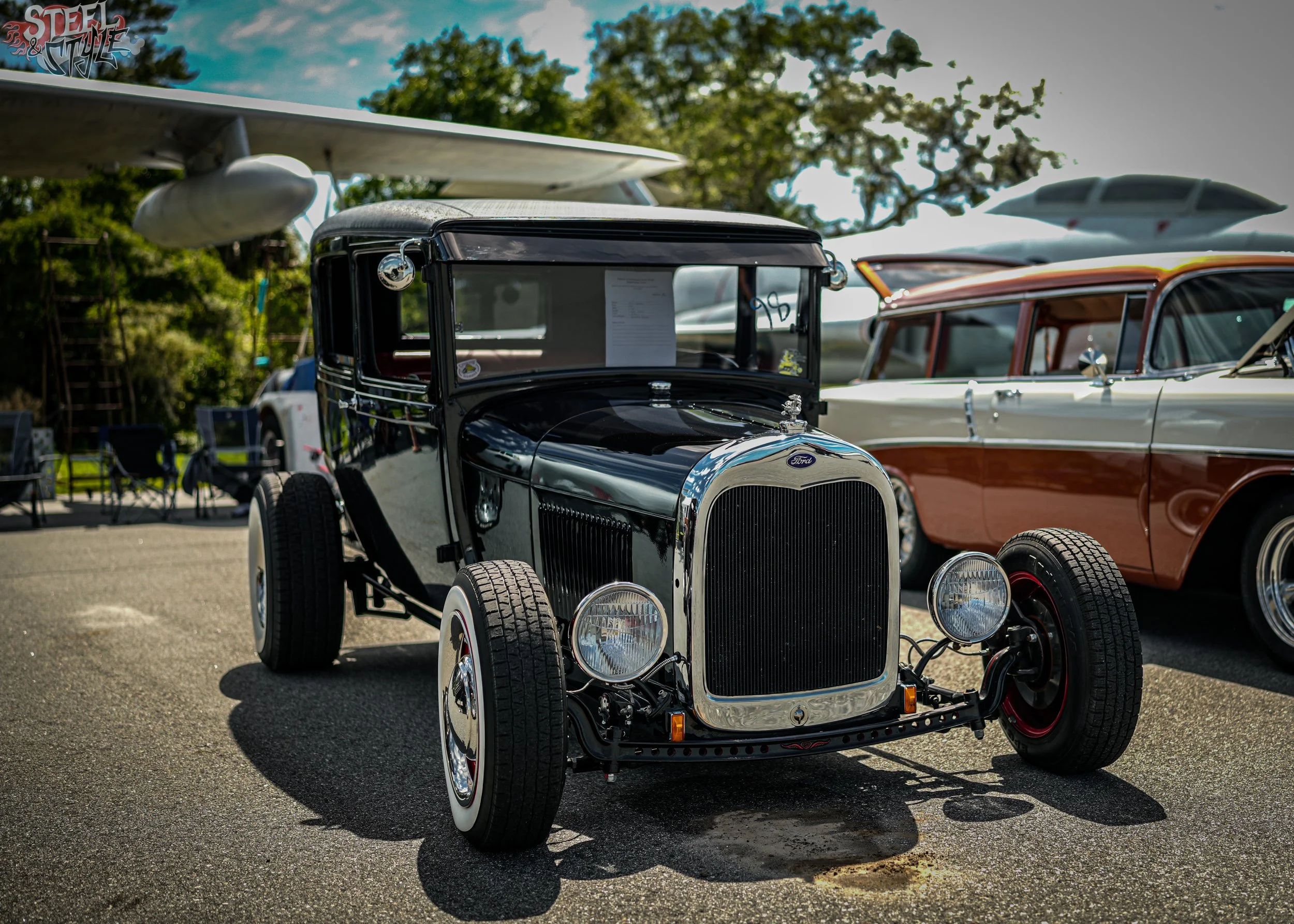 A vintage black Ford Model T car with white wall tires displayed at an outdoor car show, with aircraft models in the background and a 