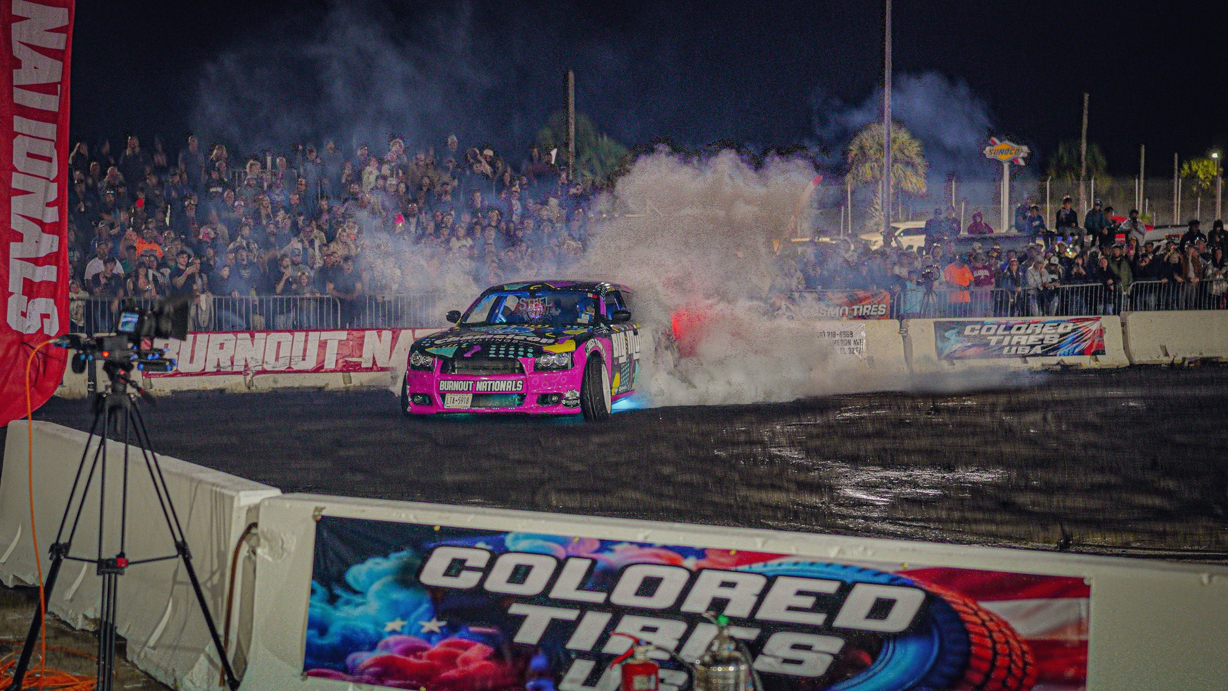 A brightly colored race car spinning on a dirt track with dust and smoke billowing behind it during a night race, with a large crowd of spectators watching behind a barrier.