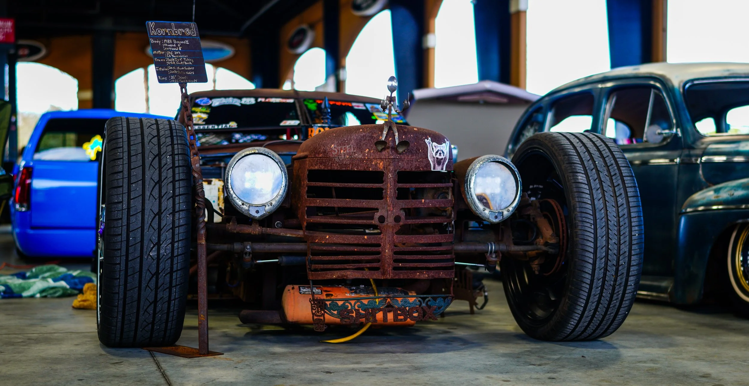 Rusty vintage car with large tires and headlamps, displayed in an indoor warehouse with other classic cars and items in the background.