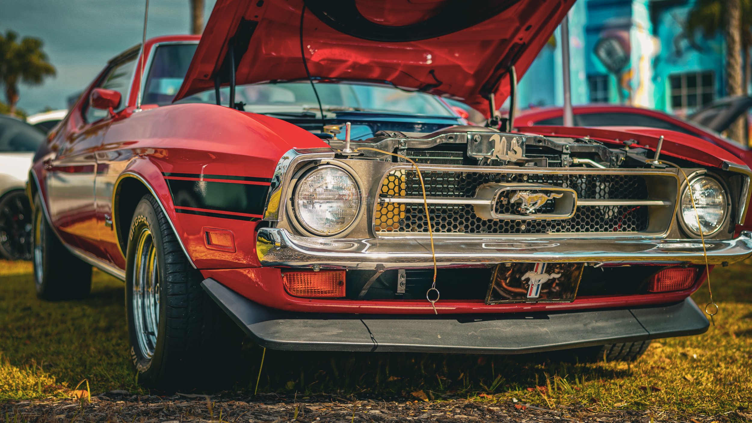A red vintage Ford Mustang car with its hood open and a partially disassembled front grille, displayed at an outdoor car show.