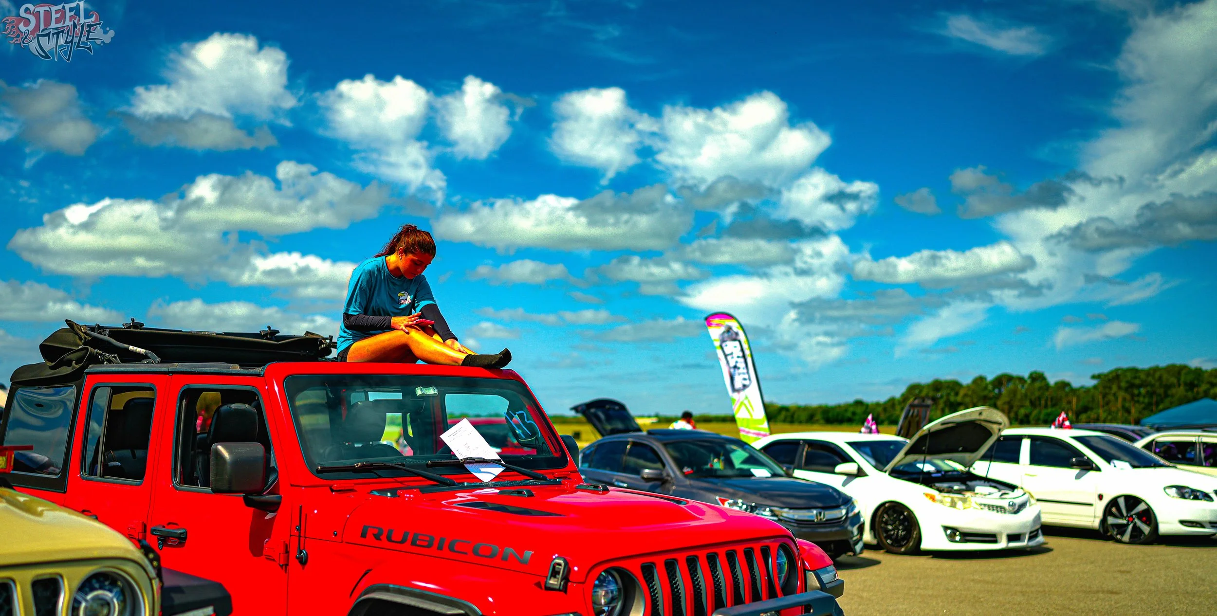 A woman sitting on top of a red Jeep Rubicon with other cars parked behind her under a partly cloudy blue sky.