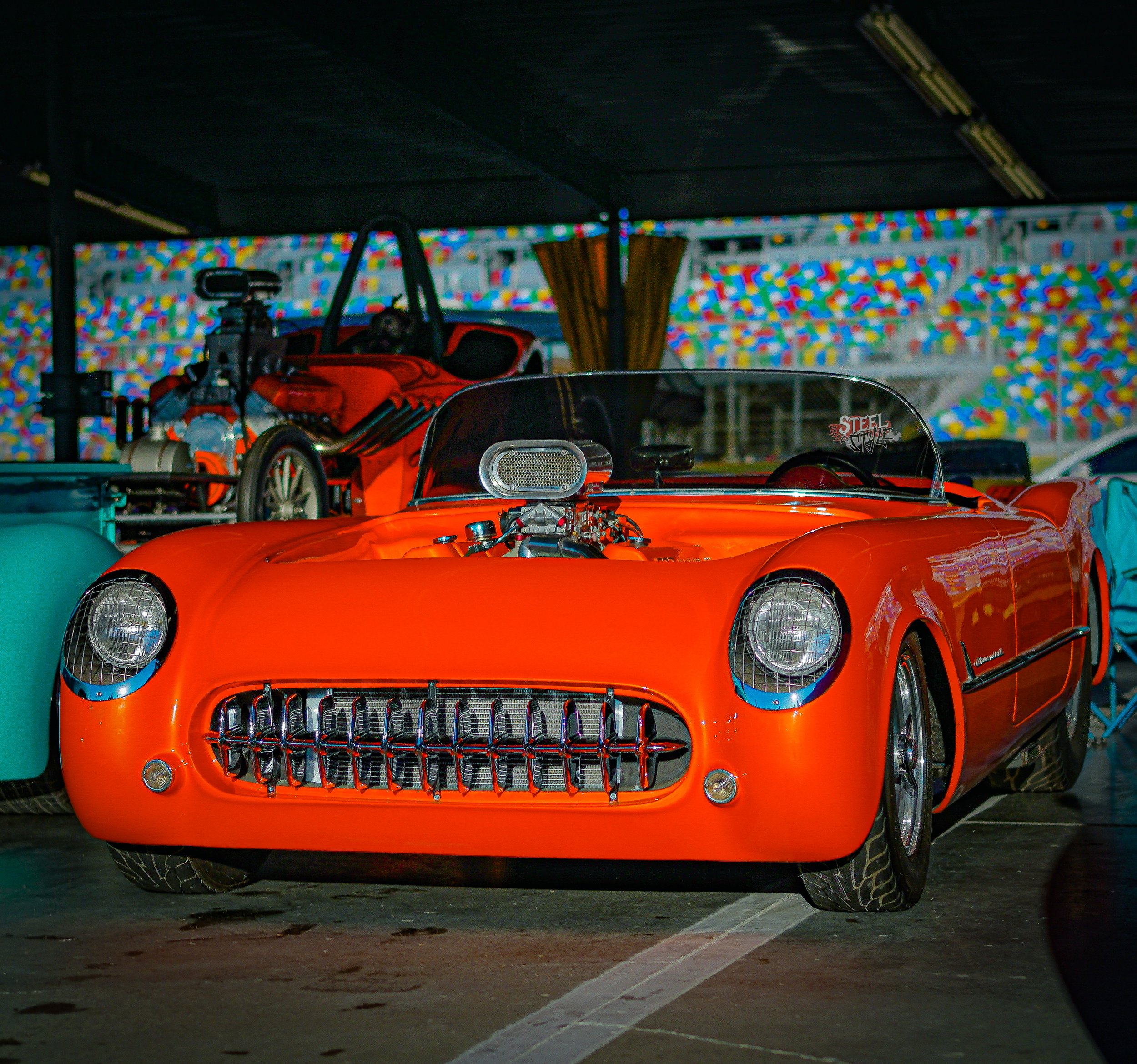 A bright orange vintage race car with a distinctive grille and exposed engine, parked indoors at a car show.