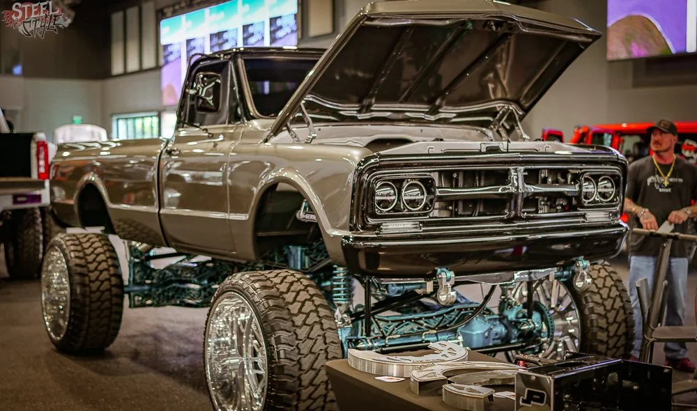 A modified gray pickup truck with large off-road tires on display at an indoor auto show, with its hood open revealing the engine.