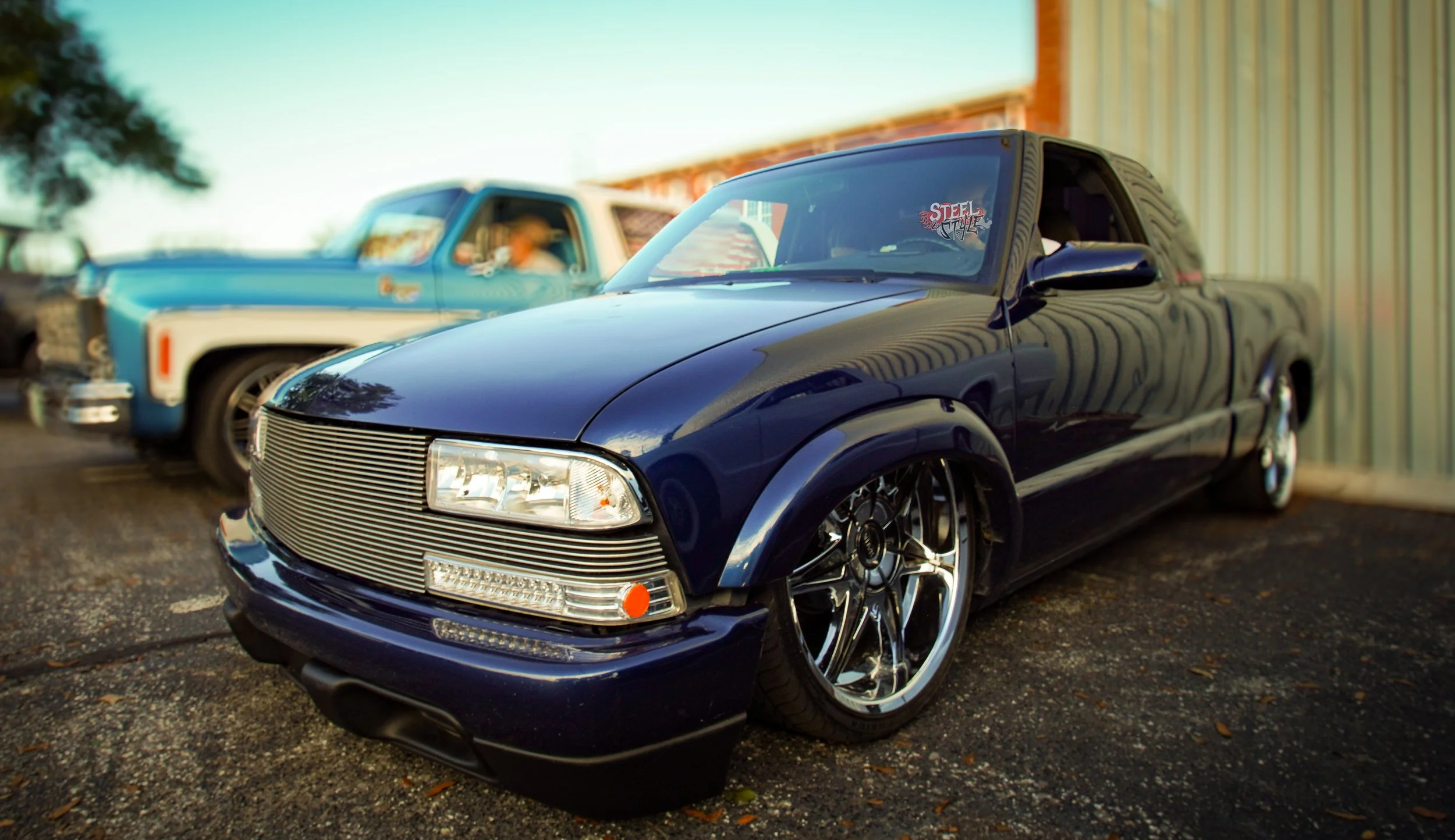 Black modified car with custom grille and wheels parked outdoors, with a blue truck and a metal building in the background.