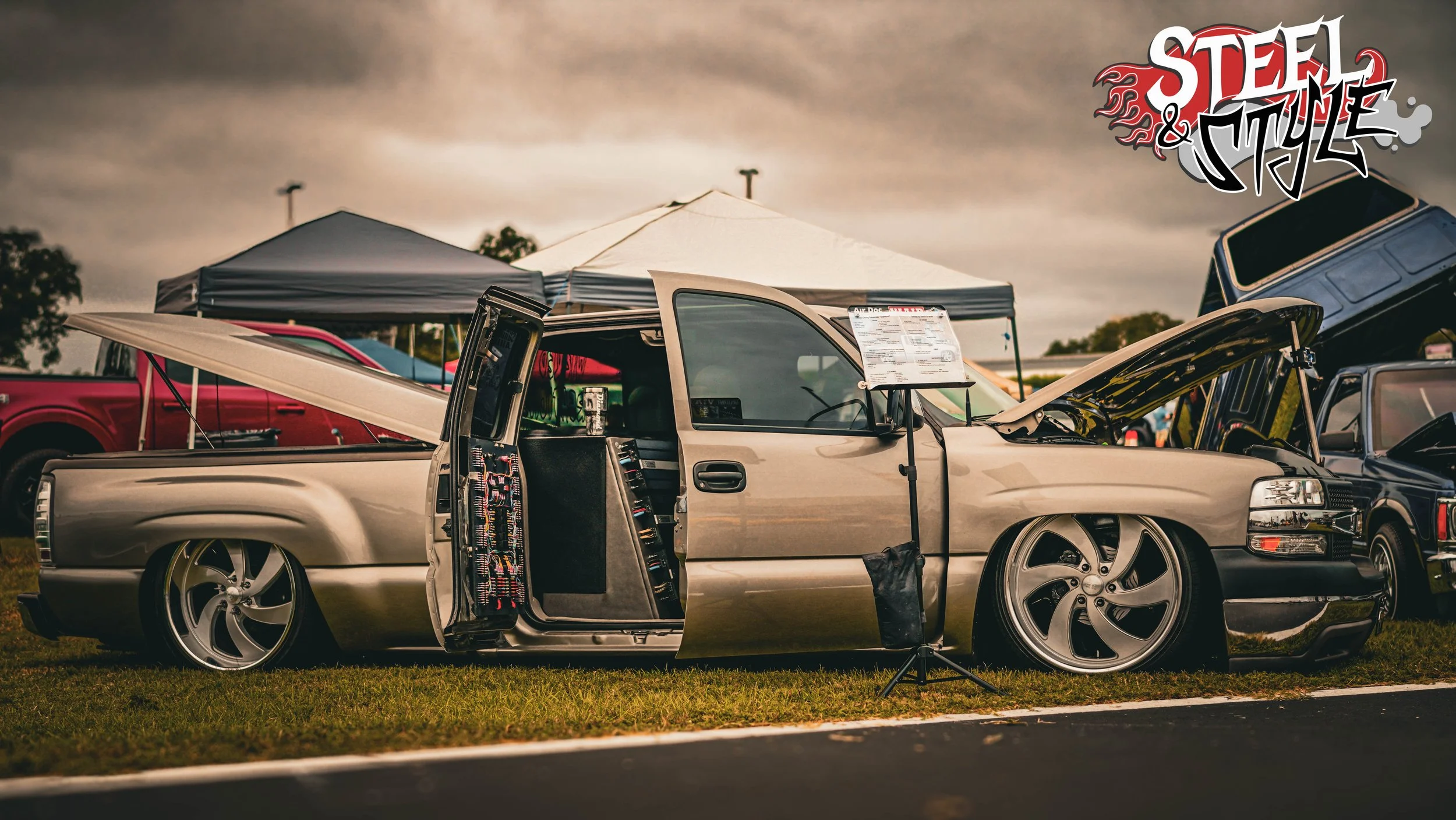 A lowered, customized beige pickup truck with open hood and open side panel at a car show, with tents and other vehicles visible in the background, and a graffiti-style "Steel & Style" logo in the top right corner.