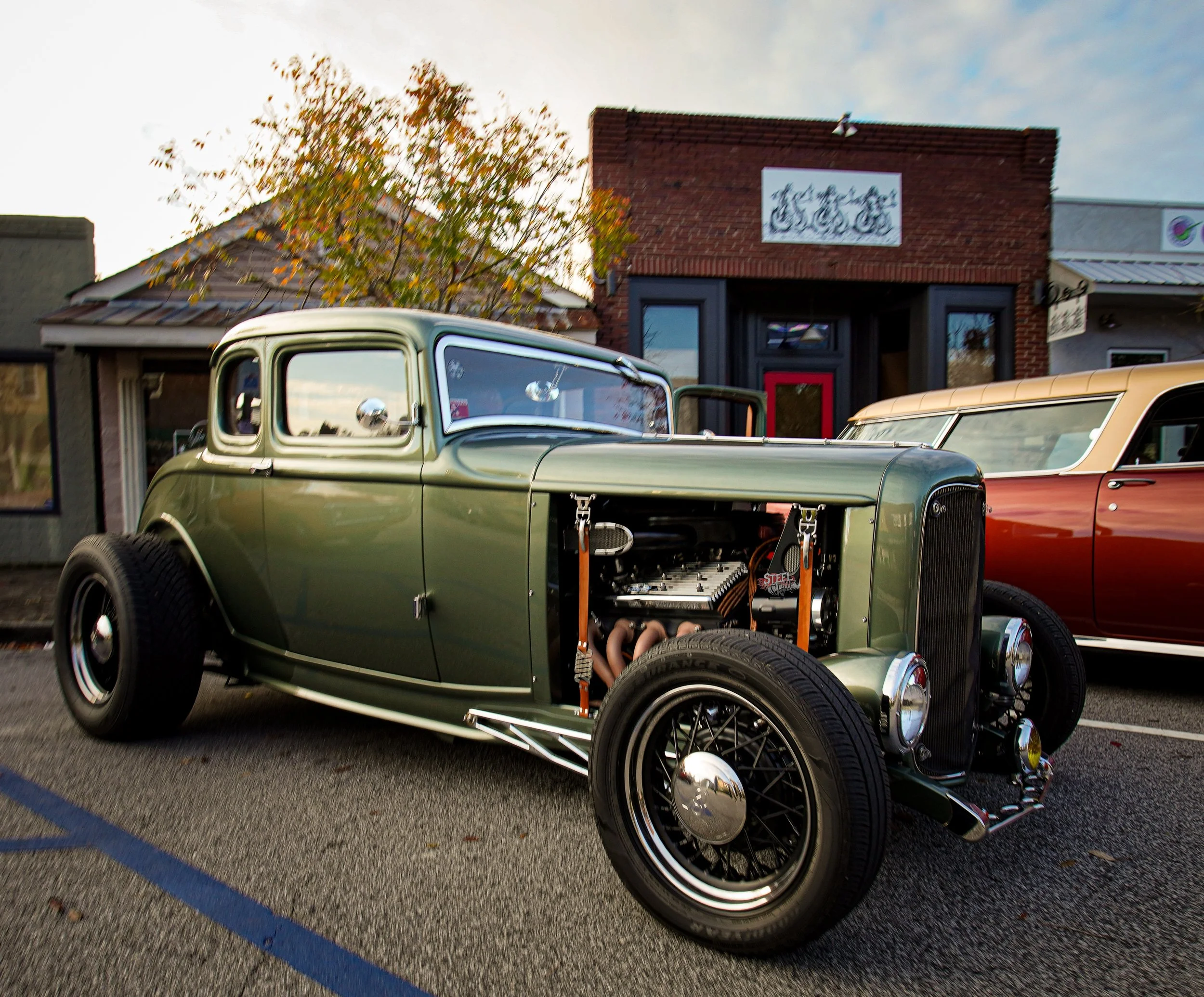 A vintage green hot rod car with exposed engine, parked on a city street during daylight, alongside other classic cars, with buildings and trees in the background.