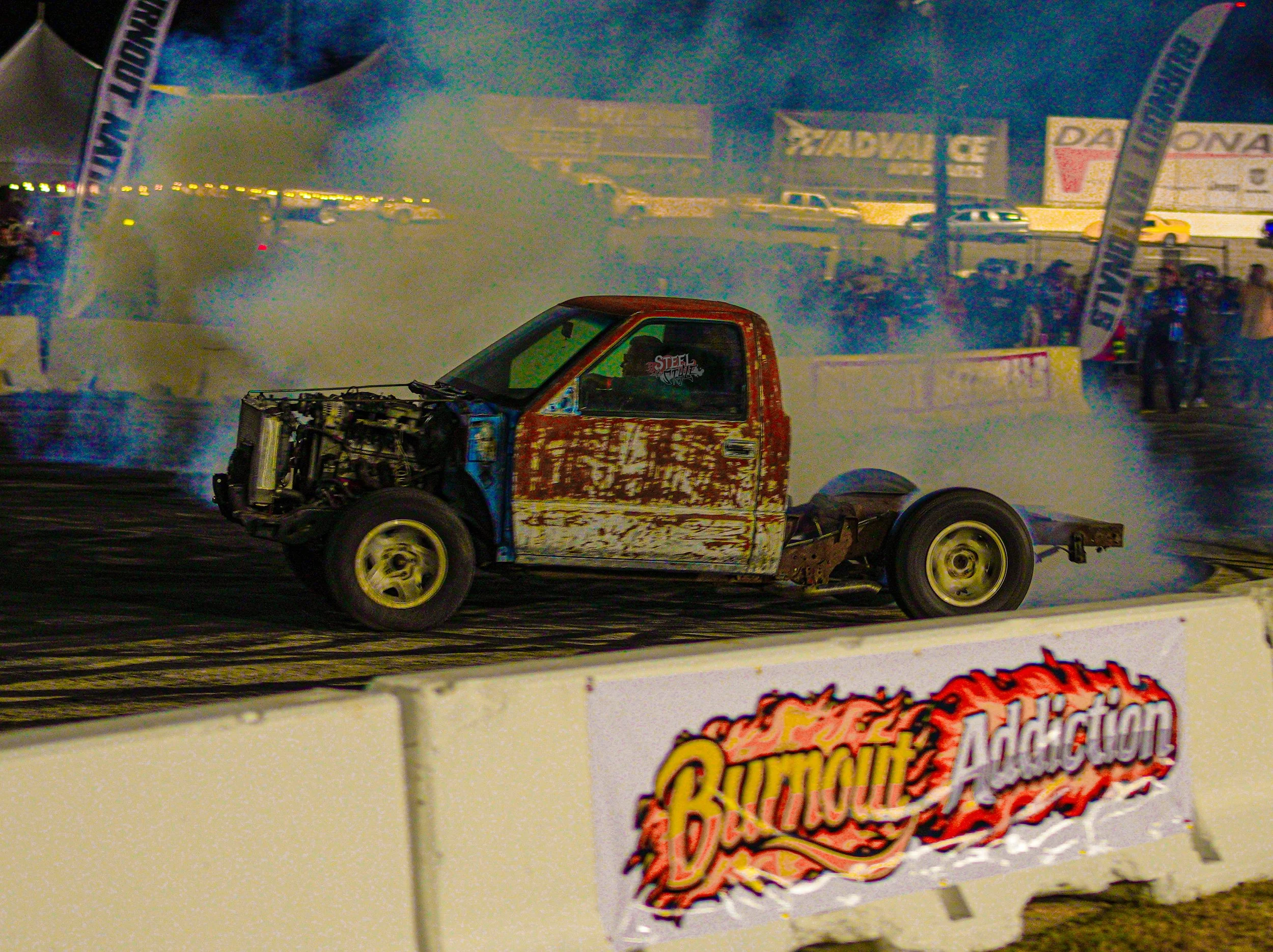 A race car with a rusted and worn exterior performing a burnout at a drag racing event, with smoke billowing around it and spectators in the background.