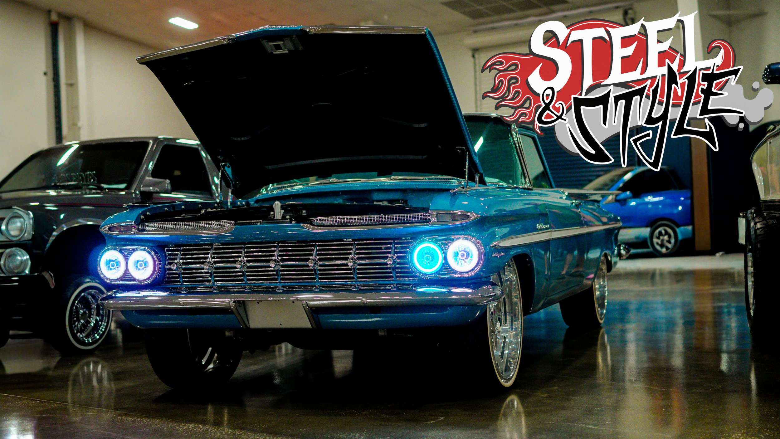 A classic blue car with its hood open, illuminated headlights, and chrome wheels, displayed indoors at a car show, with graffiti-style text reading 'Steel & Style' in the top right corner.