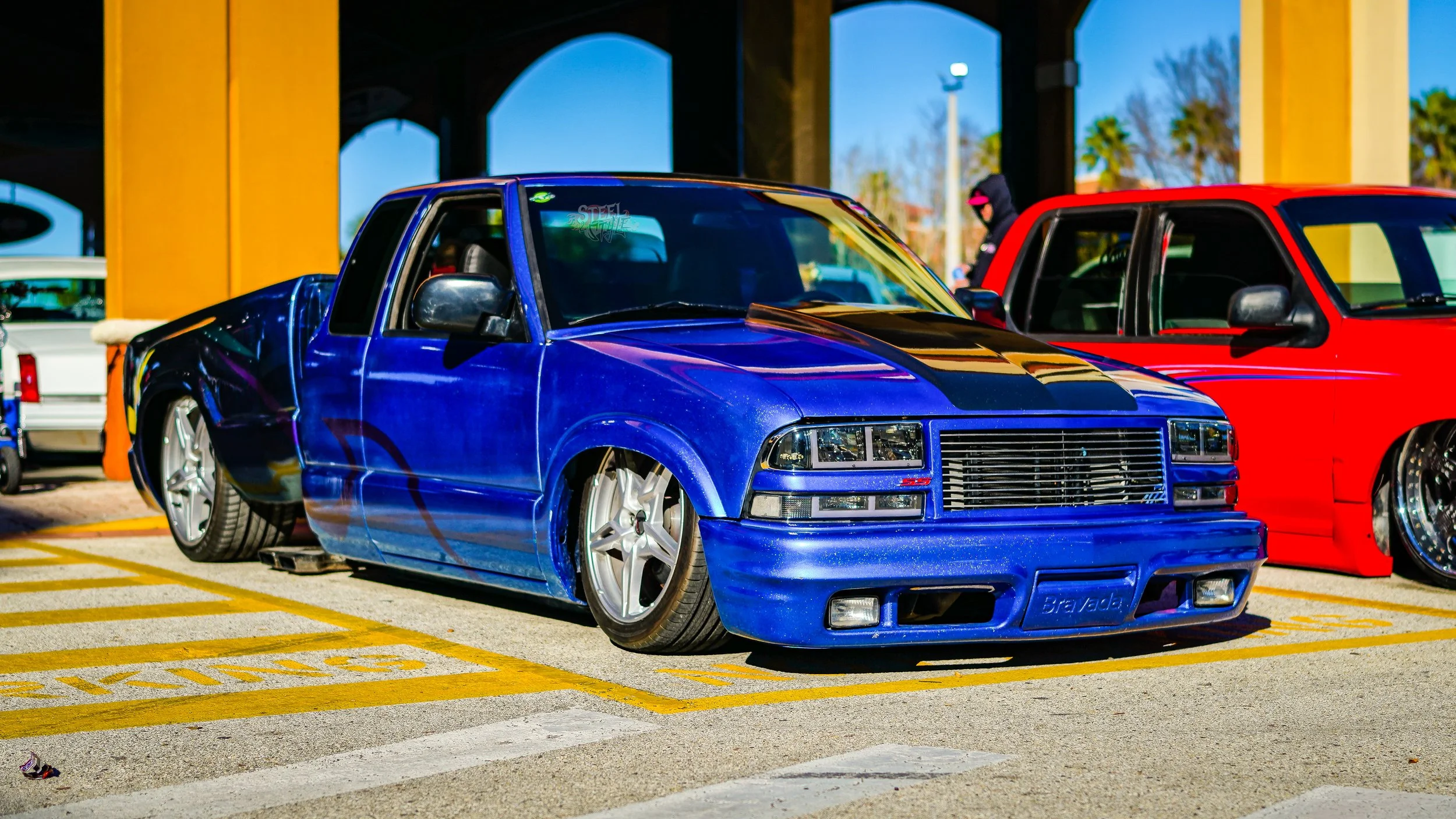 Blue modified pickup truck with black racing stripe parked in a yellow marked parking space at a car show, with other vehicles and people in the background.