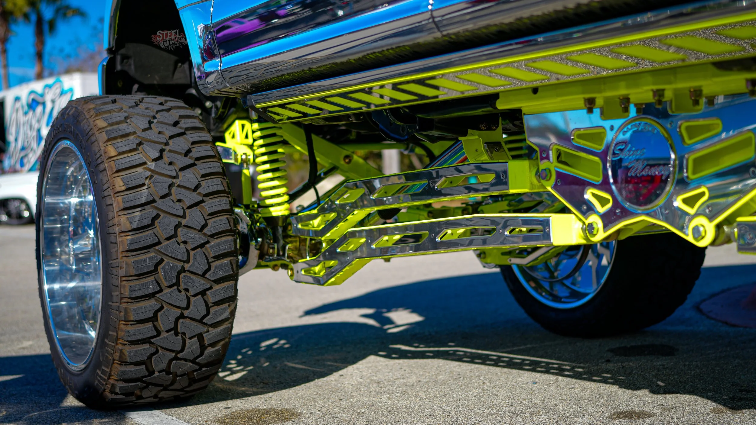Close-up photo of a customized, lifted truck with large off-road tires, chrome details, and neon yellow-green suspension components, parked outdoors.