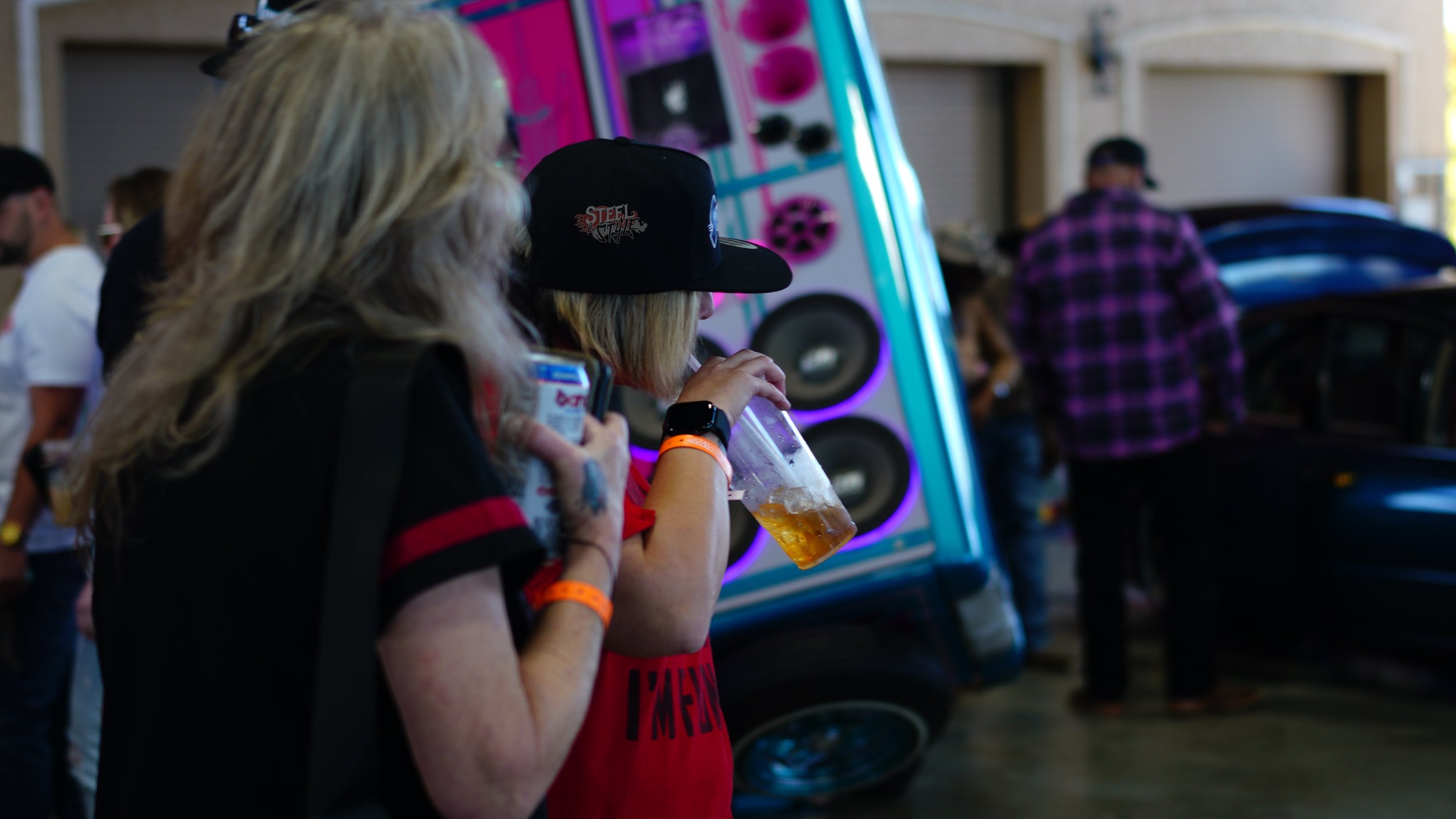 A woman with blonde hair wearing a black cap and red shirt is drinking beer from a clear plastic cup at a crowded indoor event.