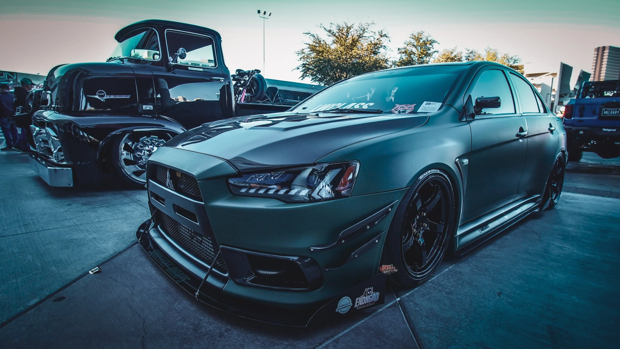 A sleek, matte green modified Nissan sedan with custom black wheels and aerodynamic body kit on display at a car show.
