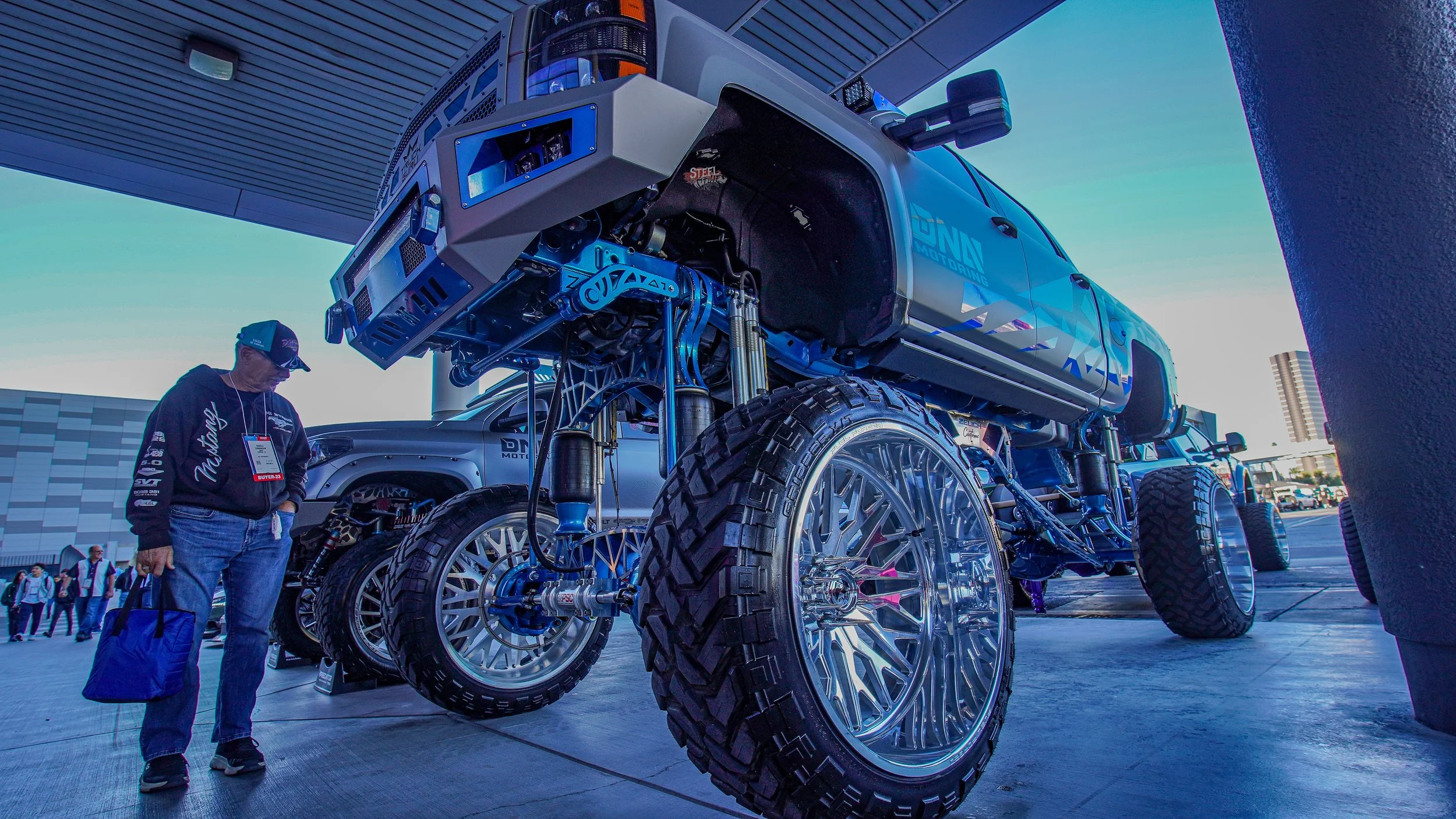 A large custom-built truck with oversized shiny chrome wheels and lifted suspension, displayed outdoors at an automotive event. A man in a black hoodie and jeans is standing nearby, looking at the vehicle.
