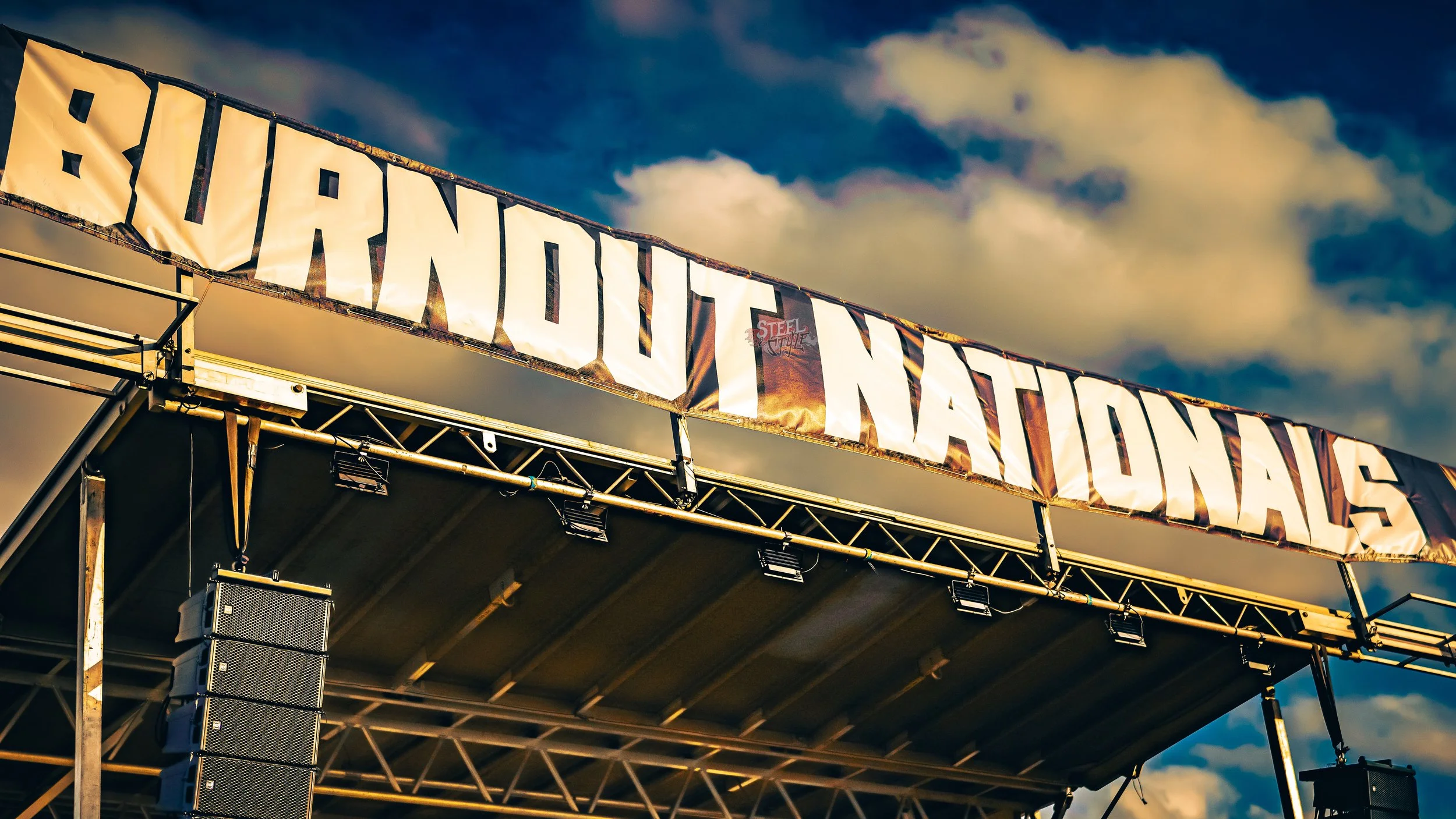 Large banner with the words 'BURNOUT NATIONALS' displayed above a stage with speakers, under a partly cloudy sky.