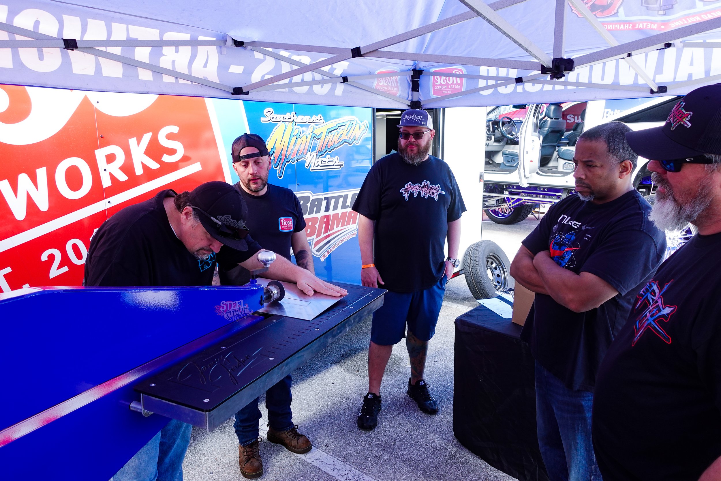 Group of men gathered under a tent at a racing event, watching a man operate a blue steel-tube machine, with racing-related banners and a vehicle in the background.