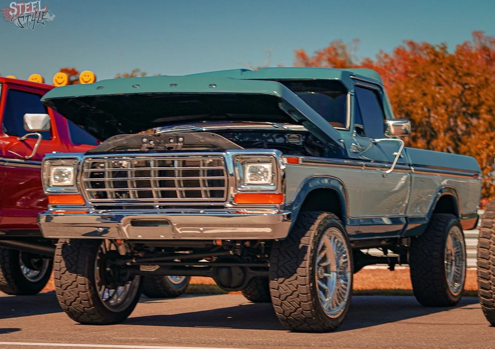 A vintage blue pickup truck with open hood, parked outdoors during daytime, with autumn trees in the background.