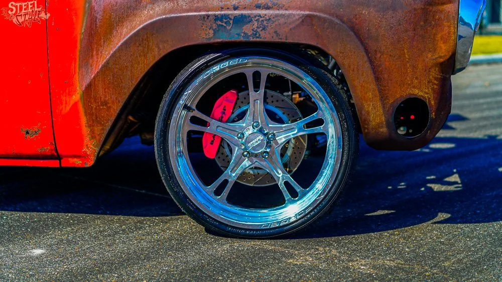 Close-up of a rusted vehicle's front wheel with a shiny, aftermarket rim and a visible red brake caliper