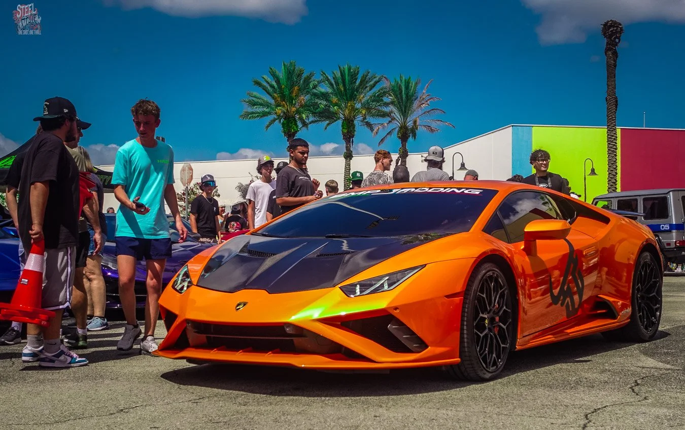 Orange Lamborghini Huracán displayed at a Dezerland Park Orlando car event, photographed from a low front three-quarter angle with spectators and palm trees in the background.