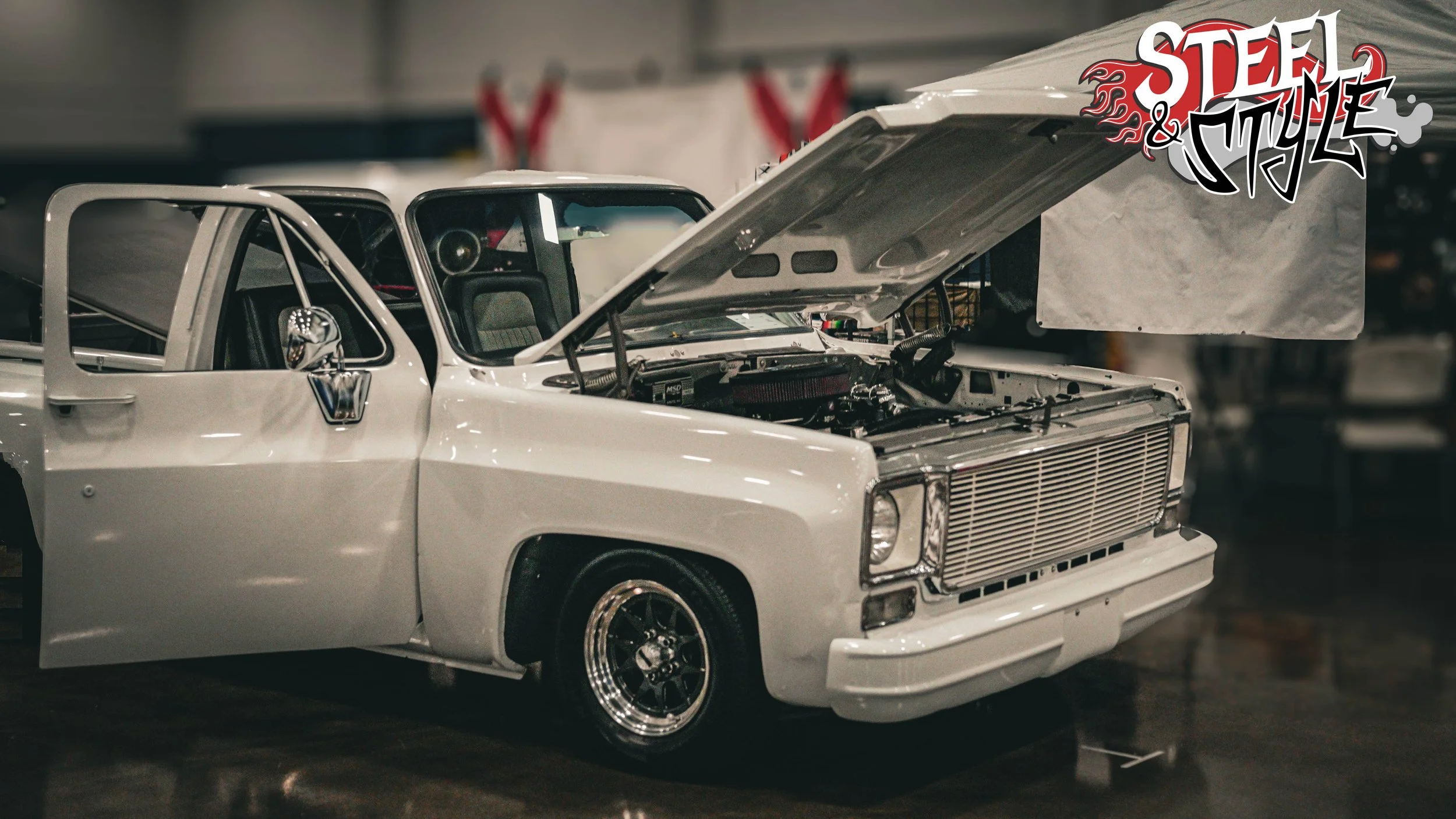 A white vintage race car with an open hood revealing the engine, displayed indoors at an event with a 'Steel & Myke' logo overlay in the top right corner.