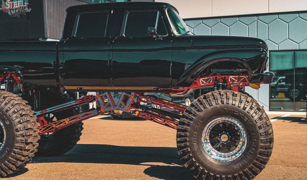 A custom-built monster truck with oversized tires and a lifted suspension, parked outside a building with glass windows.