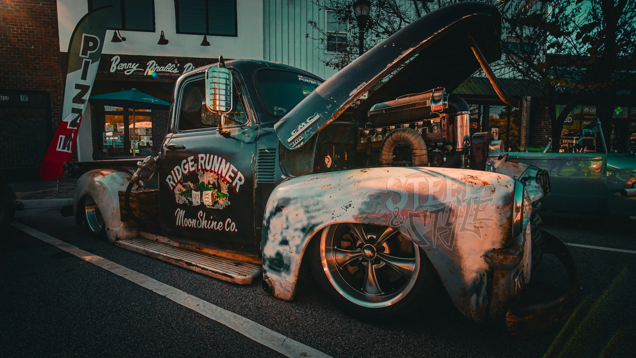 Rusty vintage pickup truck with its hood open, displaying engine, parked on street at night, with sign reading "Ridge Runner MoonShine Co." on the side, nearby storefronts and streetlamp.