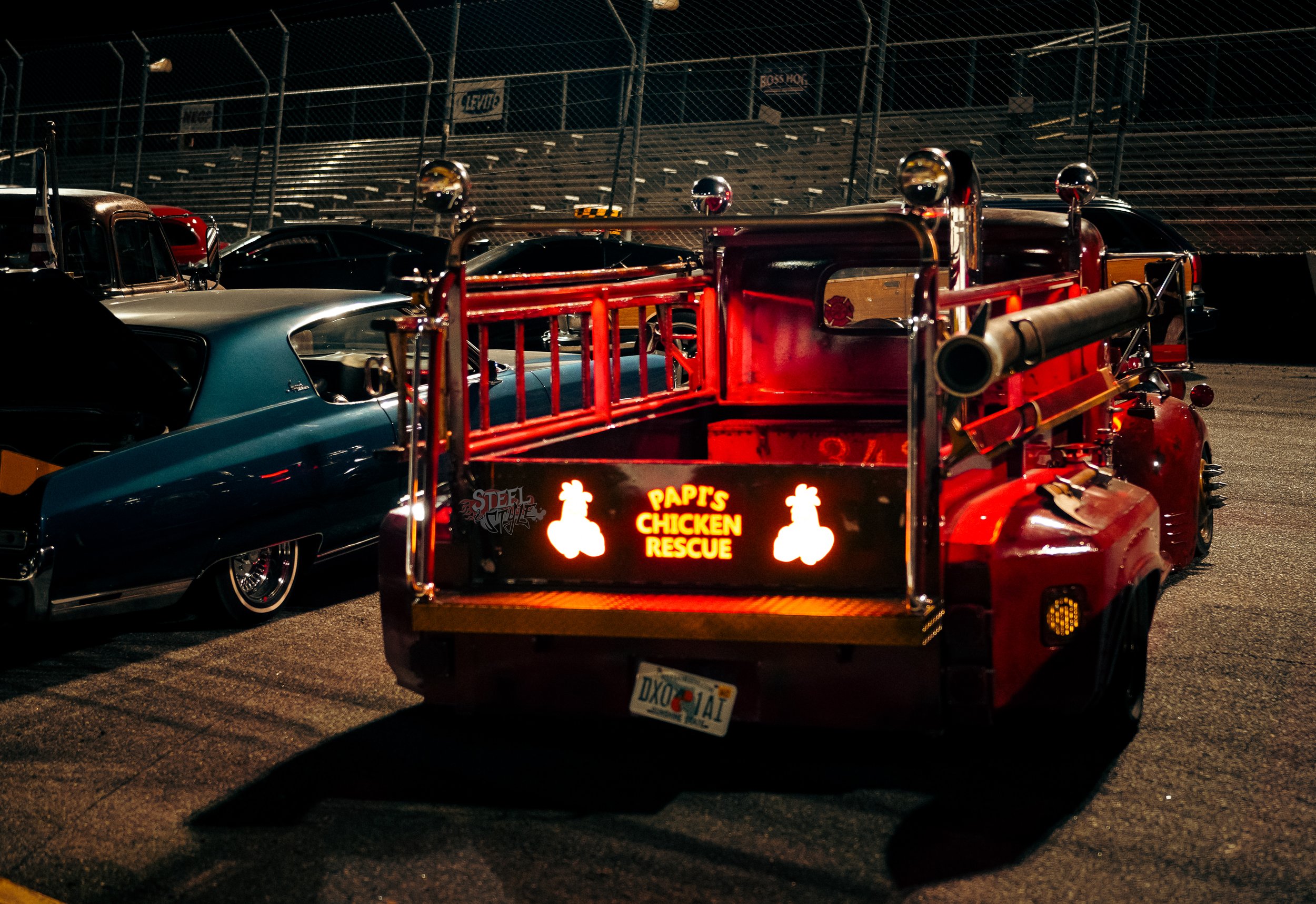 A red rescue vehicle with a sign that reads 'Pap's Chicken Rescue,' parked at a car show or event during nighttime, surrounded by other vintage cars, with a chain-link fence and bleachers in the background.