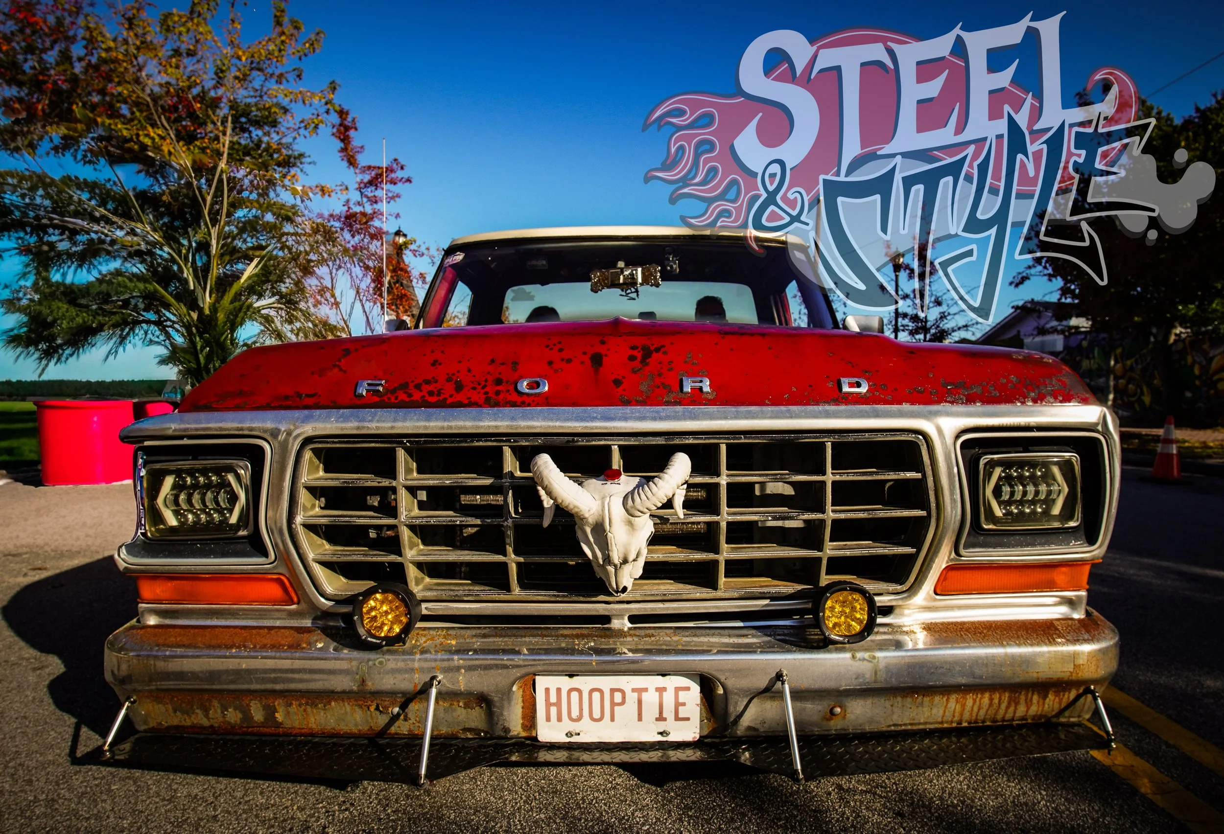 Front view of a vintage red Ford pickup truck with a bull skull mounted on the grille, a custom license plate reading 'HOOPTIE', and graffiti-style artwork that says 'STEEL & STYLE' in the background.