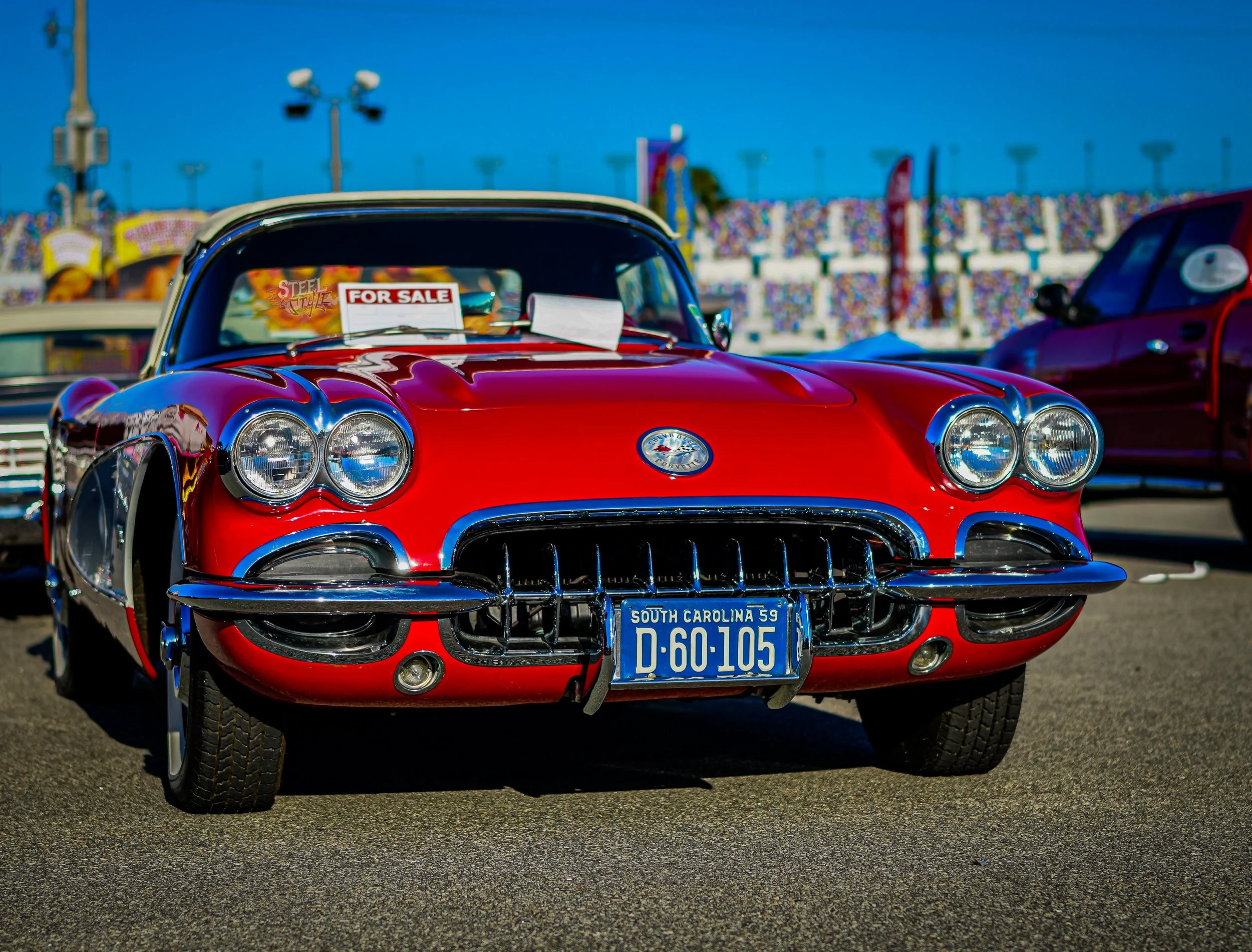 Red vintage Chevrolet Corvette with a 'For Sale' sign on the windshield, parked at a car show with other cars and a racing track in the background.