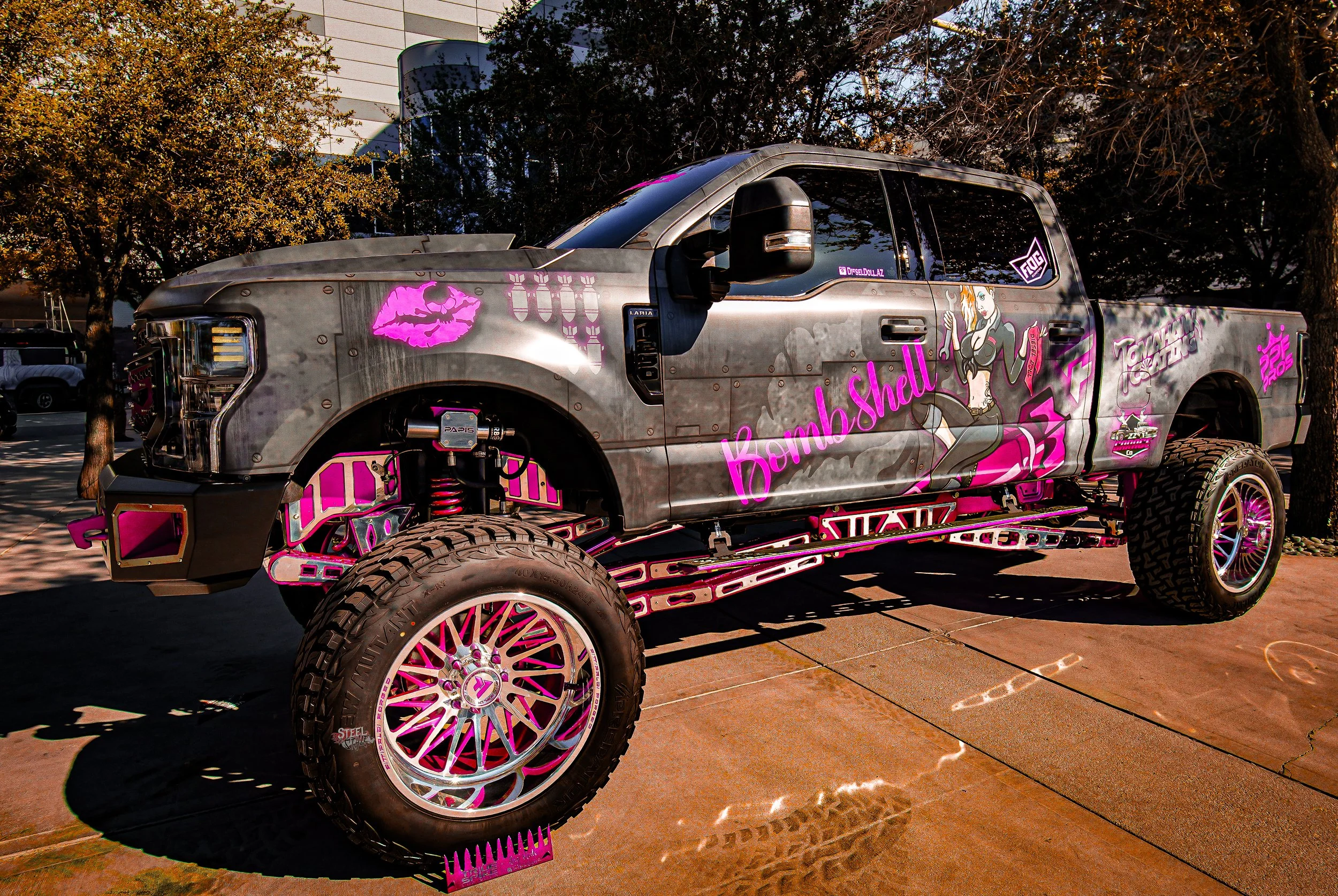A custom pickup truck with a gray and pink color scheme and graffiti-style artwork featuring women, skulls, and bombs. The side of the truck displays the words 'Bomb Shell' with a pin-up style woman holding a bomb. The truck is parked outdoors on a s