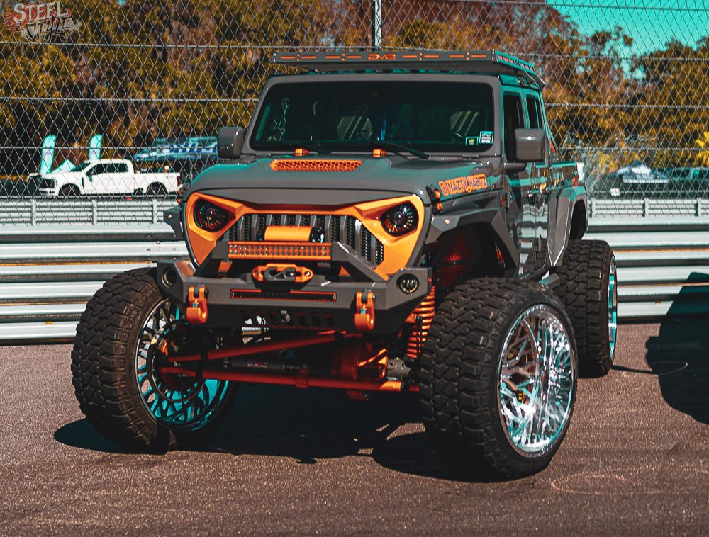 Modified off-road vehicle with oversized tires, colorful orange and black detailing, and a rugged chassis, parked in a lot with a chain-link fence and trees in the background.