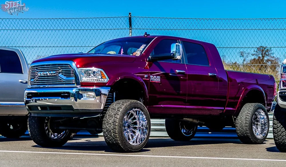 A maroon lifted Ram truck with chrome wheels parked in a parking lot next to other vehicles, with a chain-link fence and blue sky in the background.