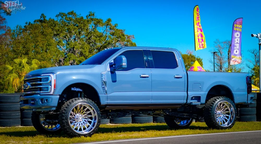A blue pickup truck with large, custom chrome wheels parked on the grass near a tire stack at an outdoor event with flags and tents in the background.