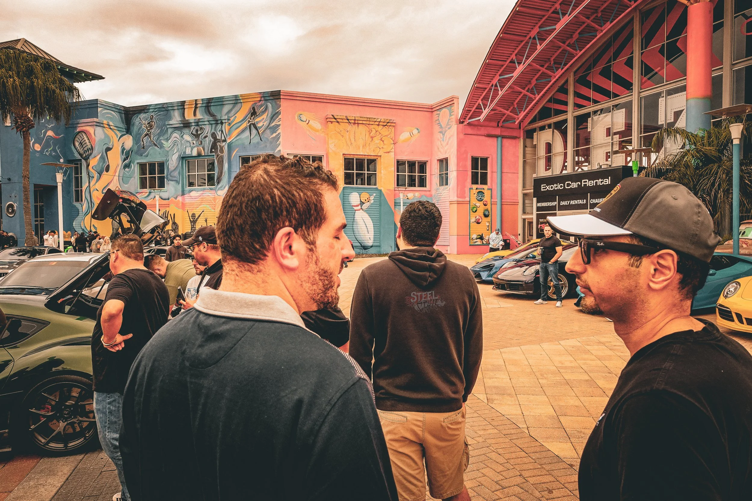 Group of people standing in front of colorful building with murals and luxury cars parked outside.