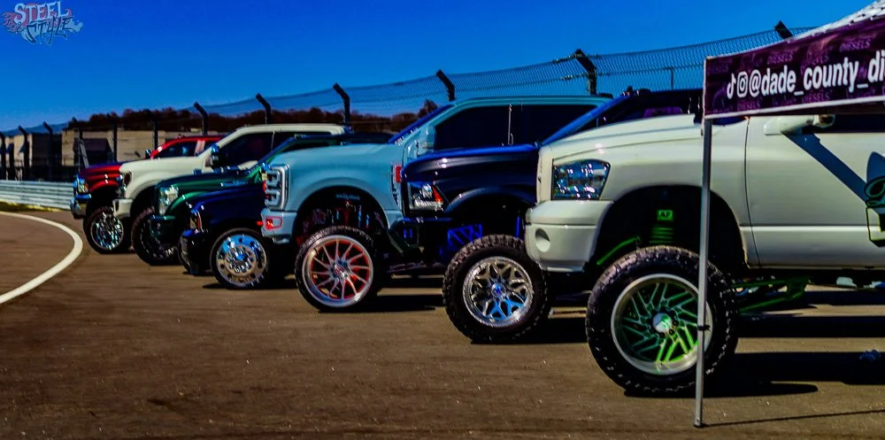 Multiple trucks lined up on a race track under a clear blue sky, with a fence and race track curving to the left in the background.